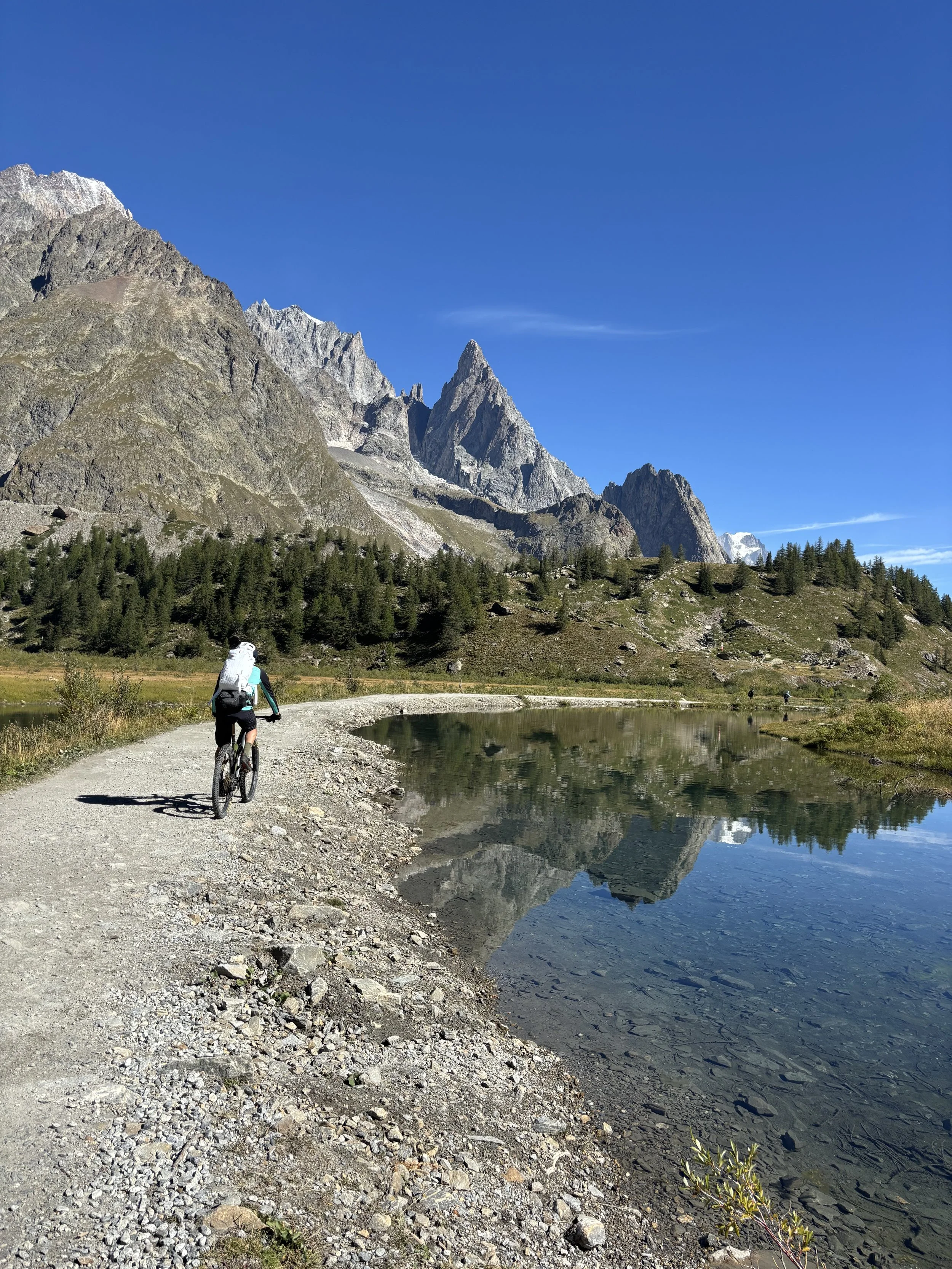 A cyclist riding on a gravel path along a mountain lake, with tall rocky peaks and a clear blue sky in the background.