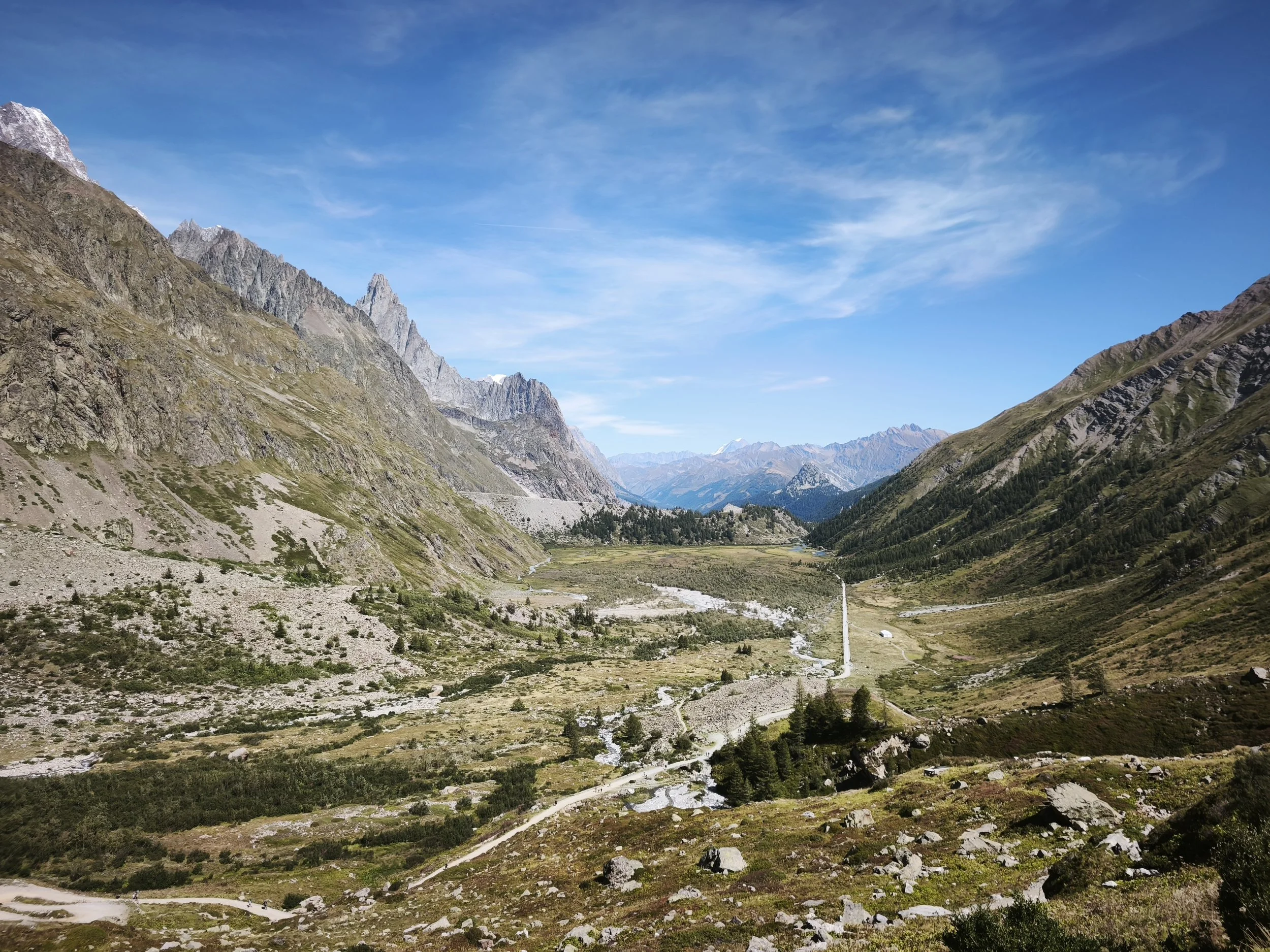 A scenic mountain valley with rugged peaks, a winding dirt road, green fields, trees, and a blue sky with wispy clouds.