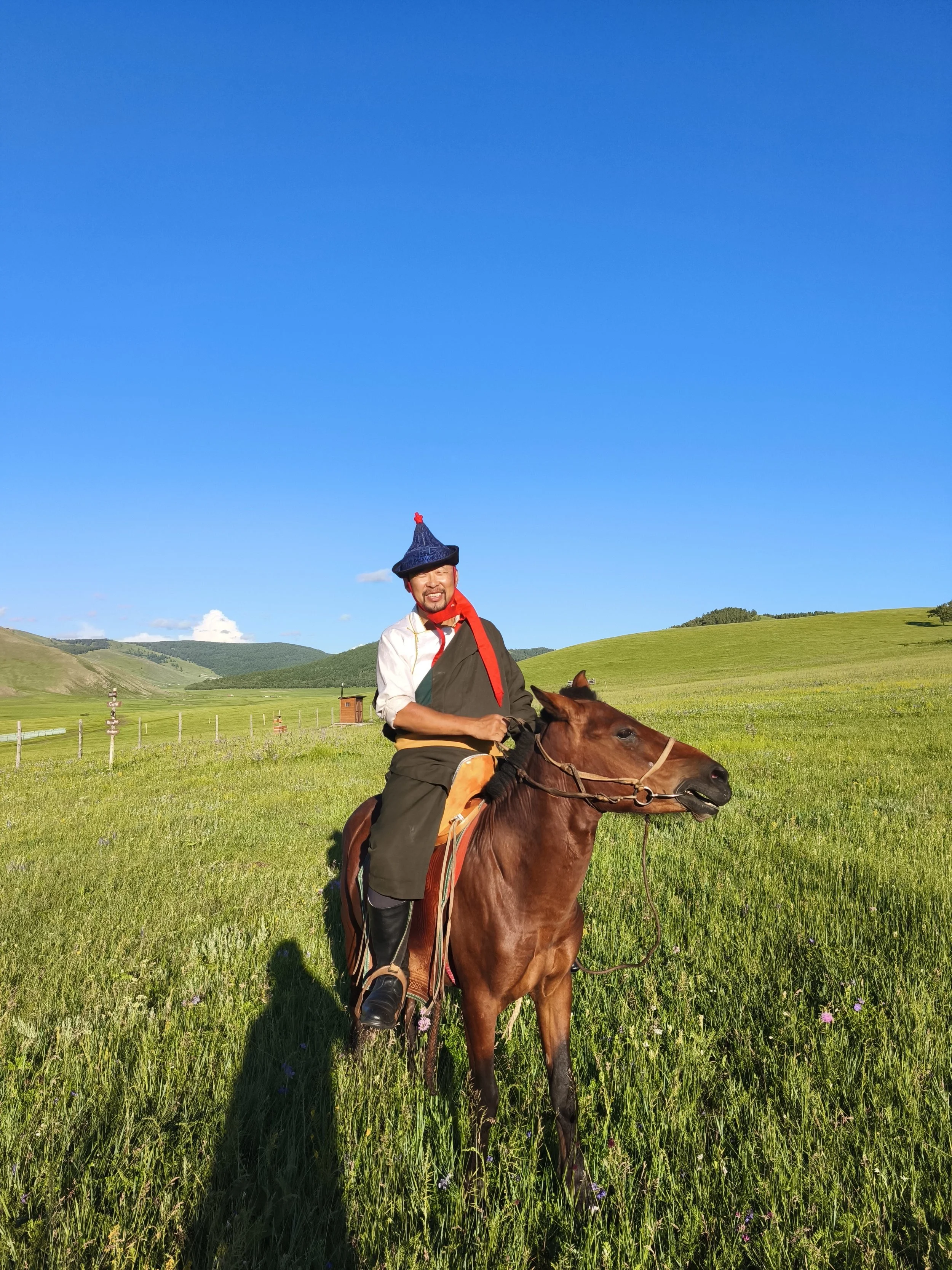 A man dressed in traditional clothing riding a brown horse in a green meadow with rolling hills under a clear blue sky.