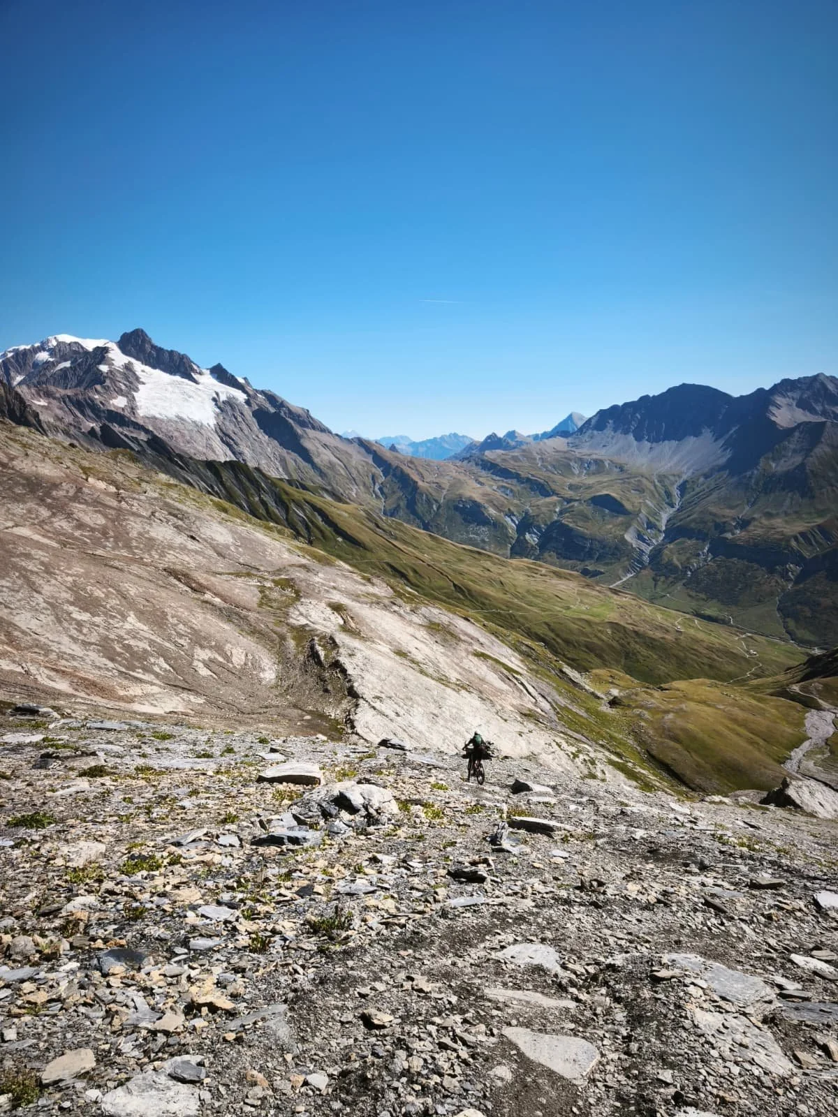 A person riding a bicycle on a rocky mountain trail with mountain peaks in the background under a clear blue sky.