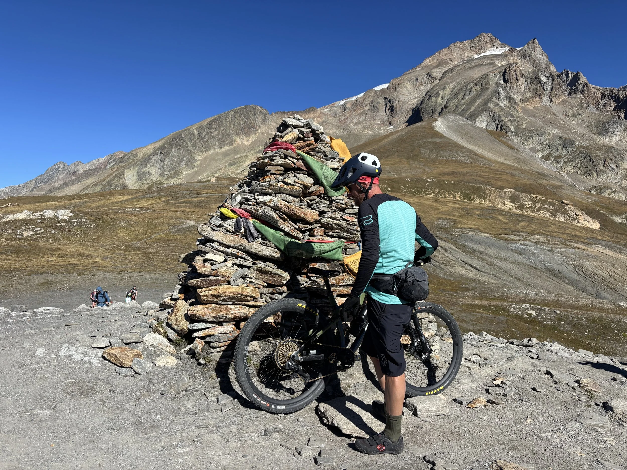 A cyclist in outdoor gear with a mountain bike, standing next to a rock cairn in a mountainous landscape with rugged peaks and clear blue sky.