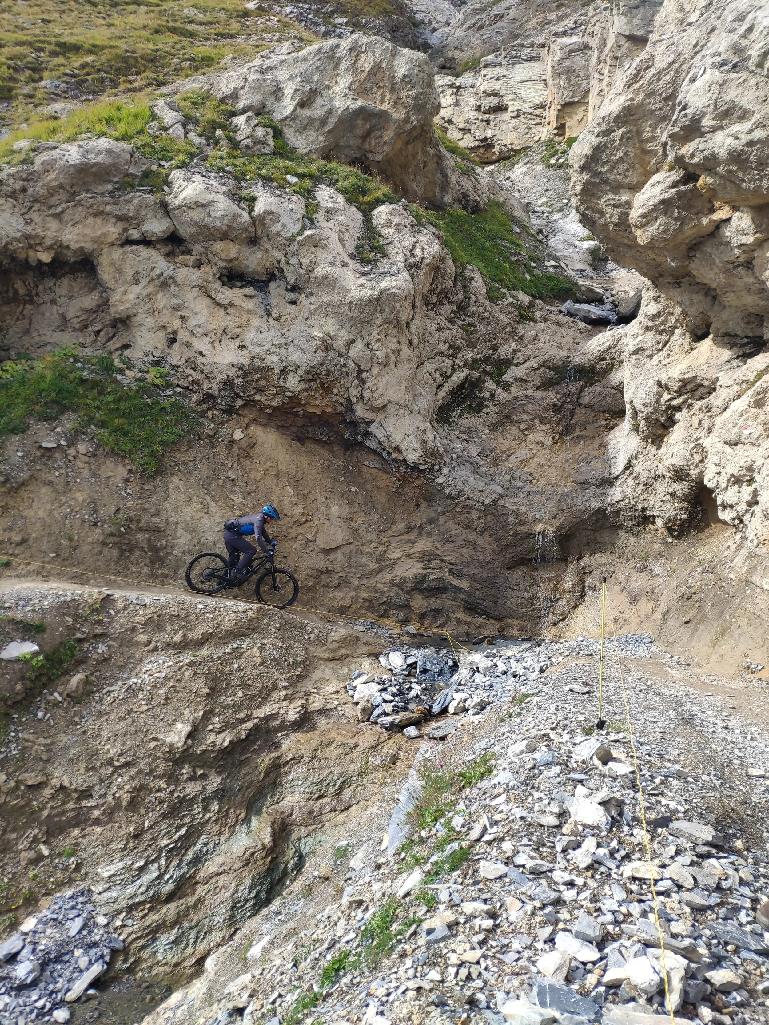 A person mountain biking on a trail in a rocky, mountainous terrain with large rocks and boulders, some green vegetation, and a steep slope.