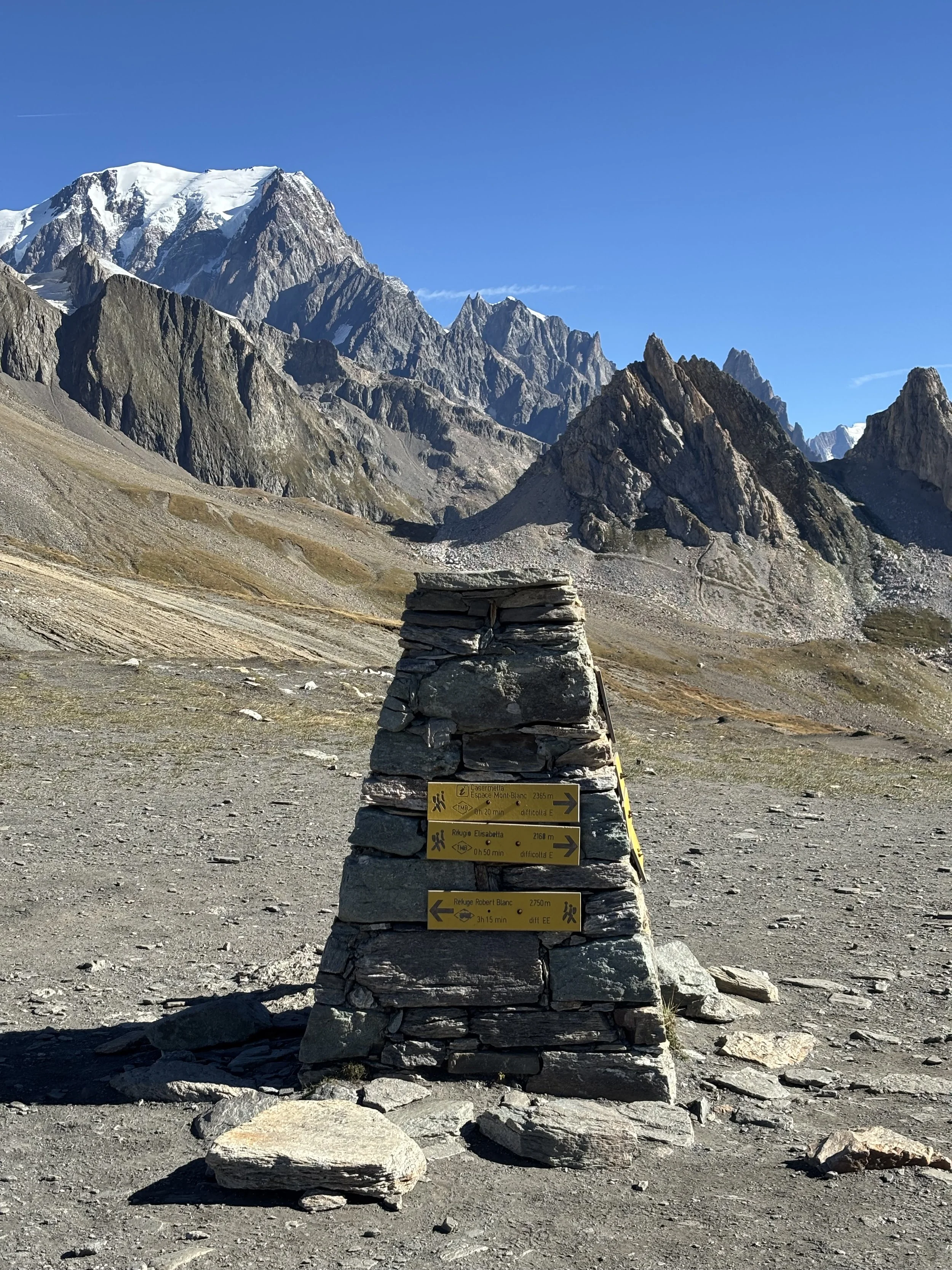 Mountain landscape with rugged peaks, some snow-capped, under a clear blue sky. Foreground features a stone cairn with yellow directional signs attached, indicating hiking routes and distances.
