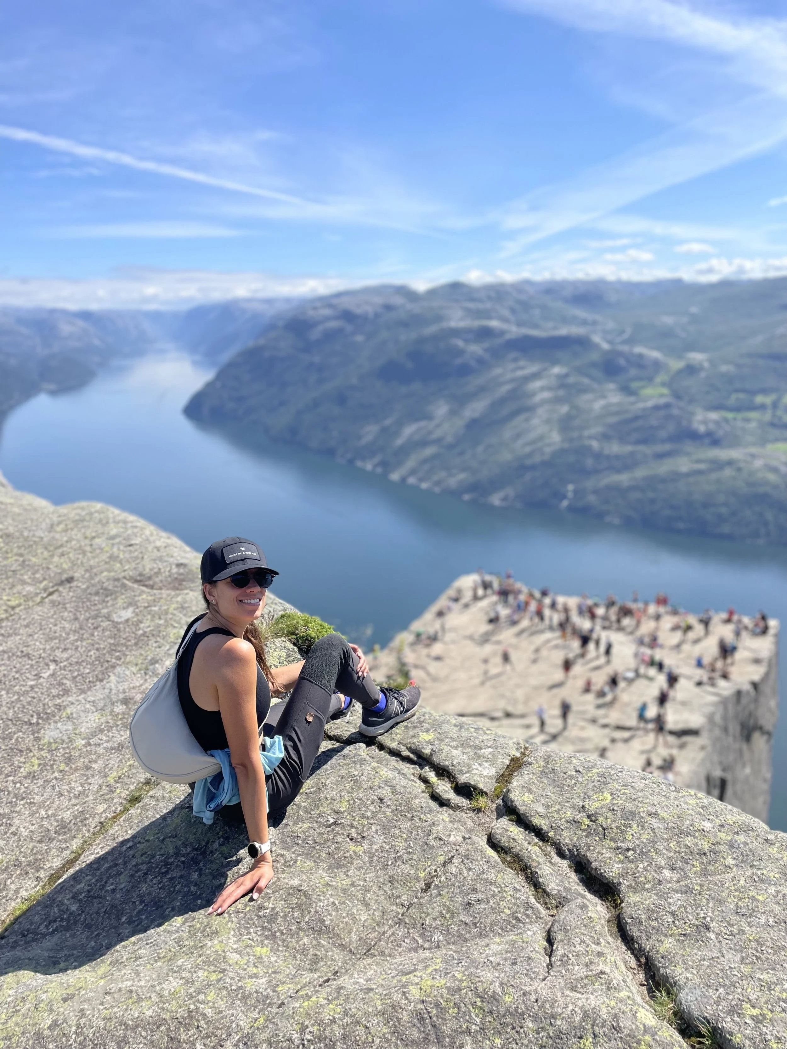 A woman with sunglasses, a black cap, and athletic attire sitting on a large rock with a scenic landscape in the background. The landscape features a river meandering through mountains under a partly cloudy sky, with many people on a hill nearby.