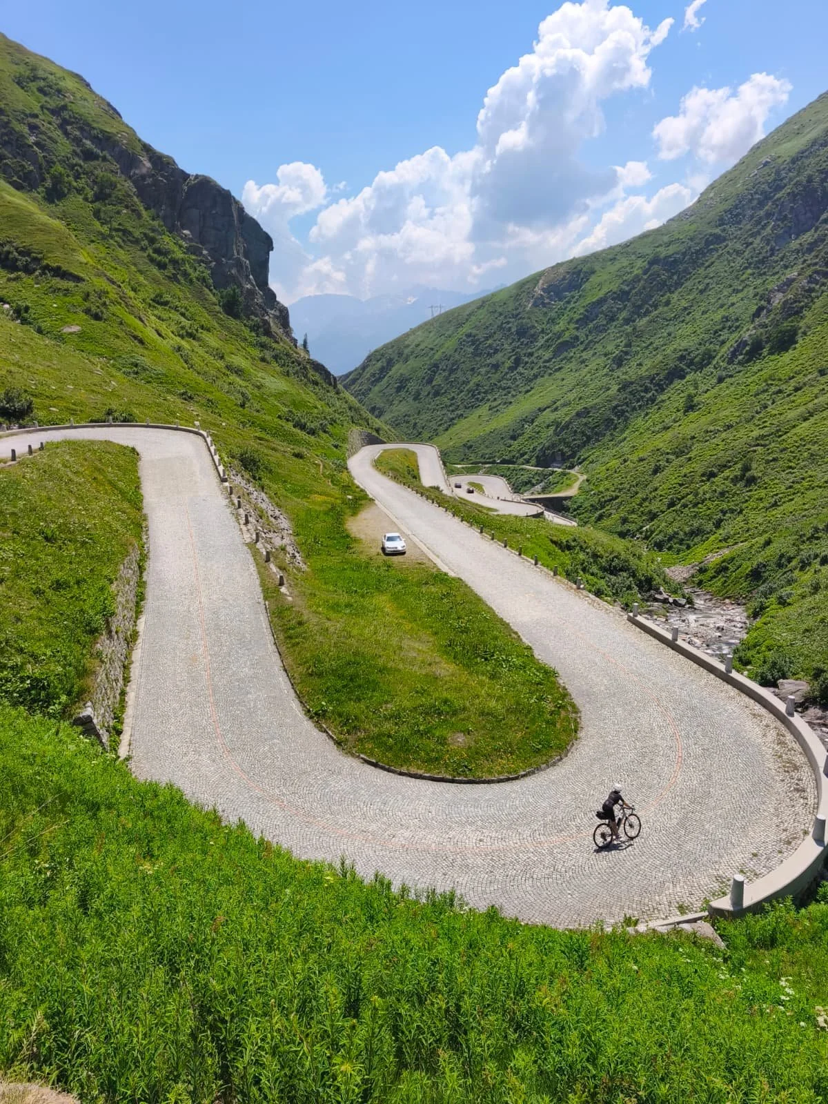 A winding mountain road with a cyclist and a parked car in a lush green valley surrounded by hills and mountains under a partly cloudy sky.