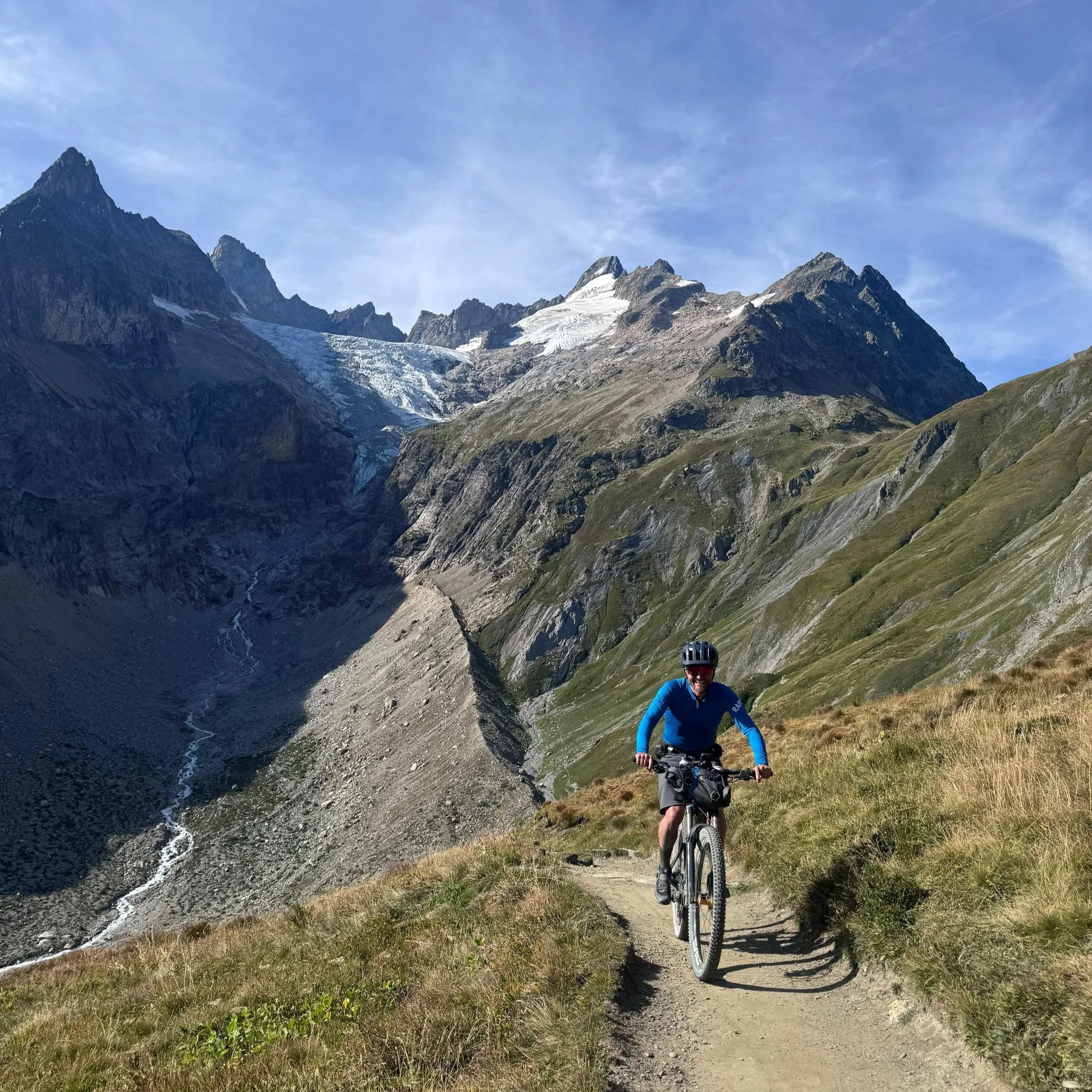 A person mountain biking on a narrow dirt trail in a mountainous area with steep slopes, rocky peaks, some patches of snow, and green grass along the trail.