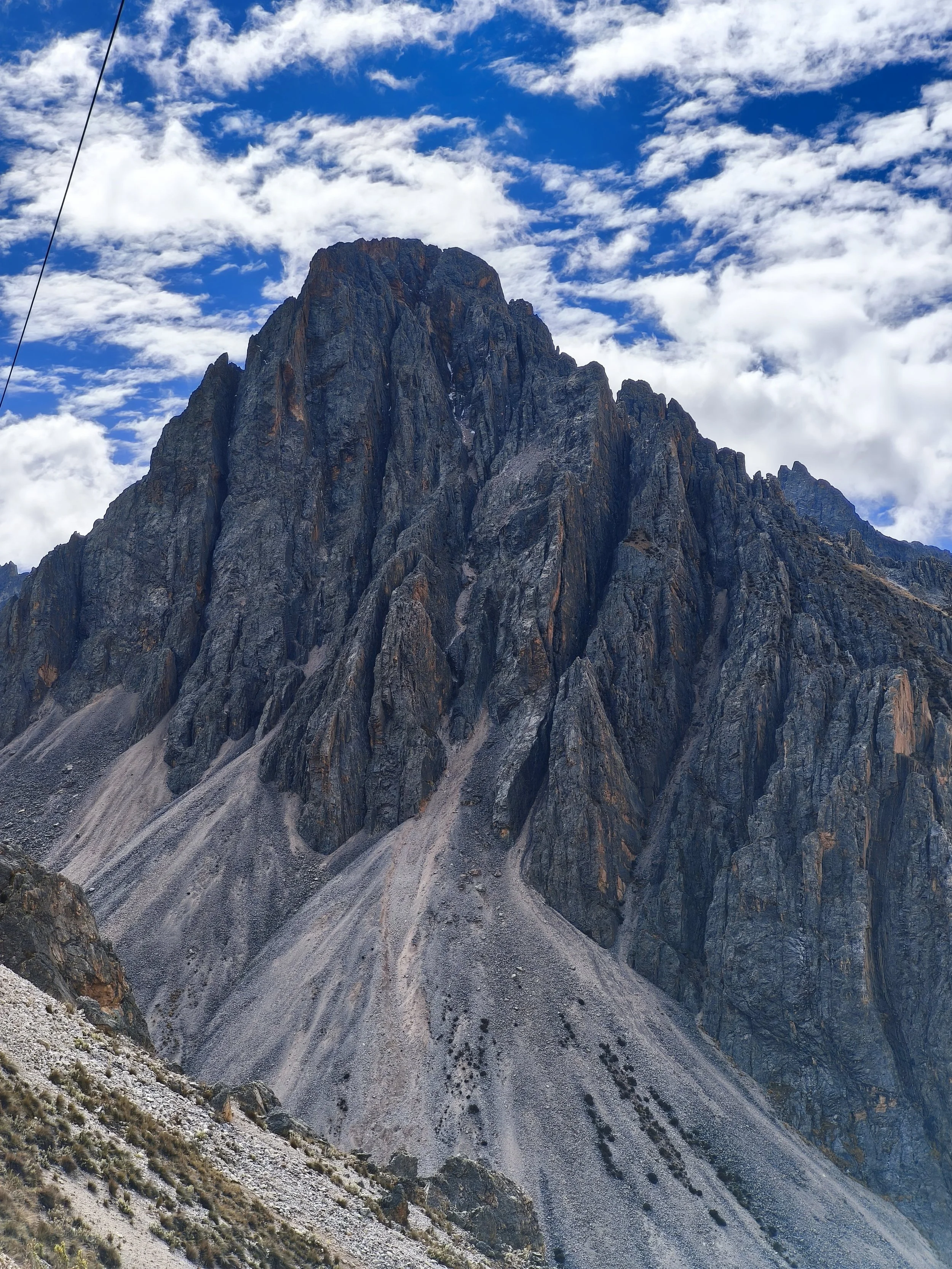 A steep mountain with dark rocky slopes and loose gravel at the base, under a partly cloudy sky with patches of blue.