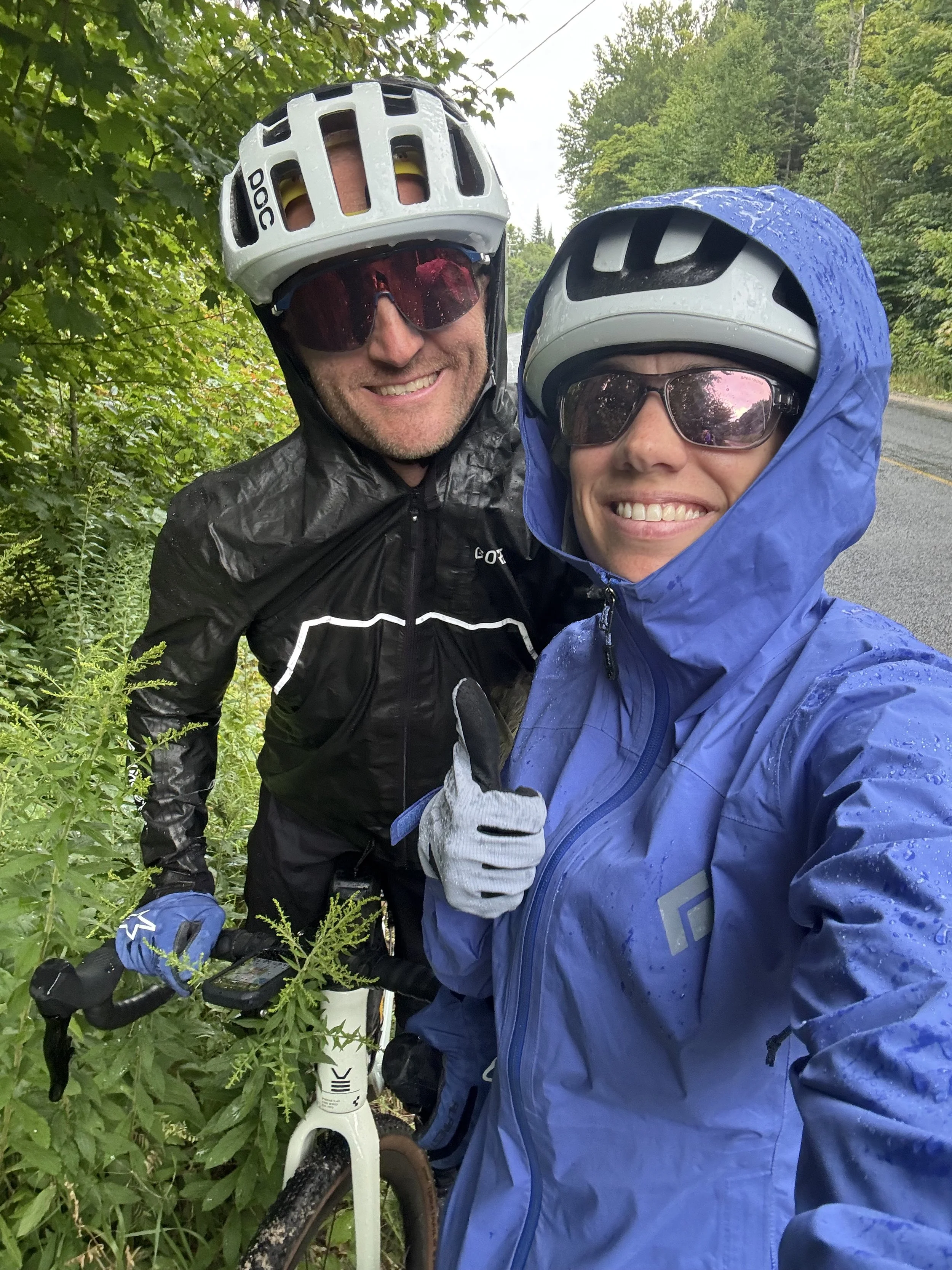 Two cyclists taking a selfie outdoors in a green, wooded area with a road in the background, wearing rain gear and helmets.