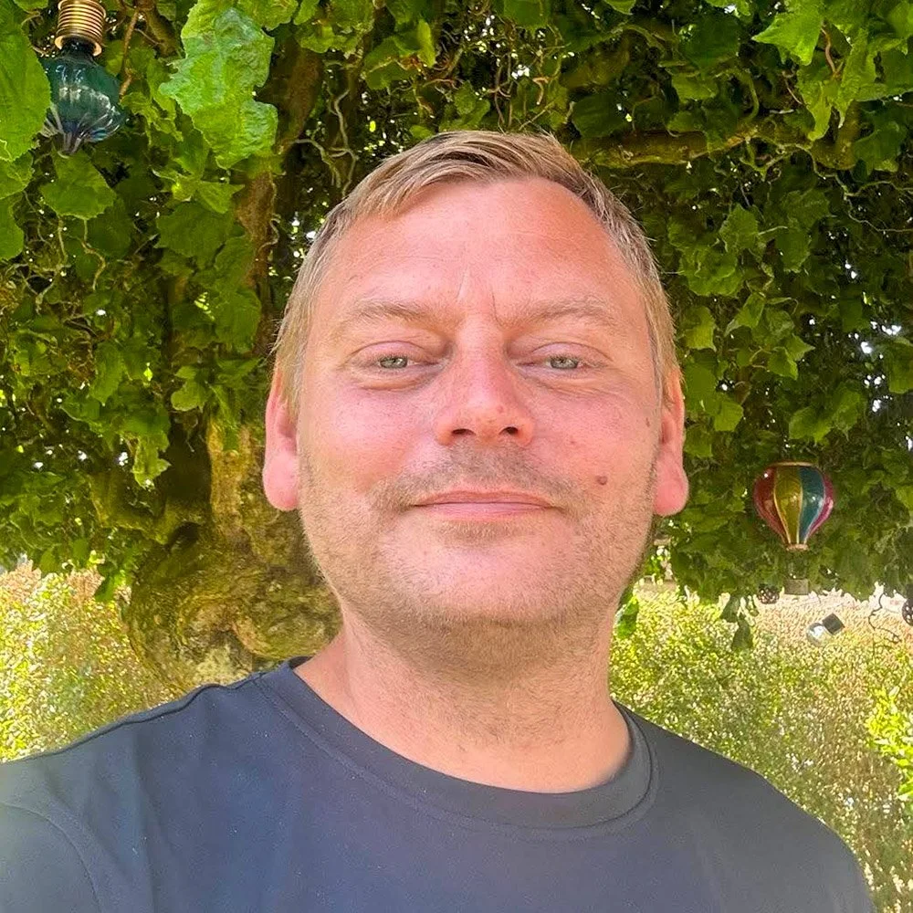Kvinde med kort blond hår, iført rustfarvet ribbet top, sidder på en bænk og læser en bog i et rum med sollys, en grøn plante og træpanel på væggen.