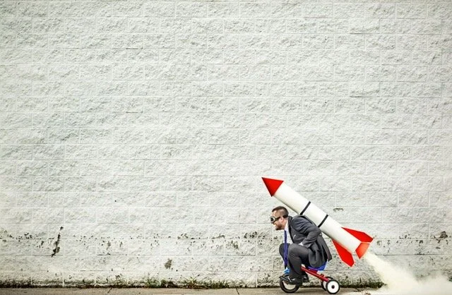Person in suit riding a scooter with a large toy rocket attached to their back, against a plain brick wall.