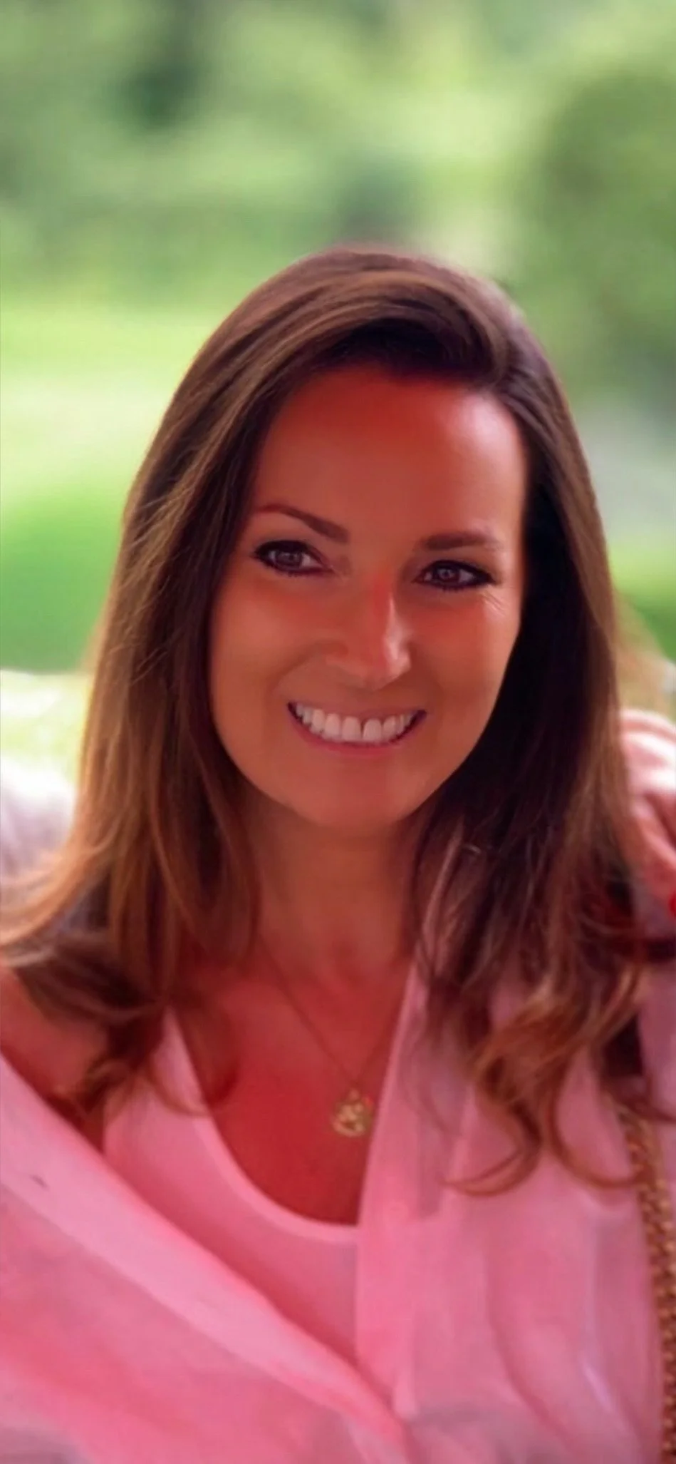 A woman with long brown hair, wearing a pink top, smiling, with a blurred green outdoor background.