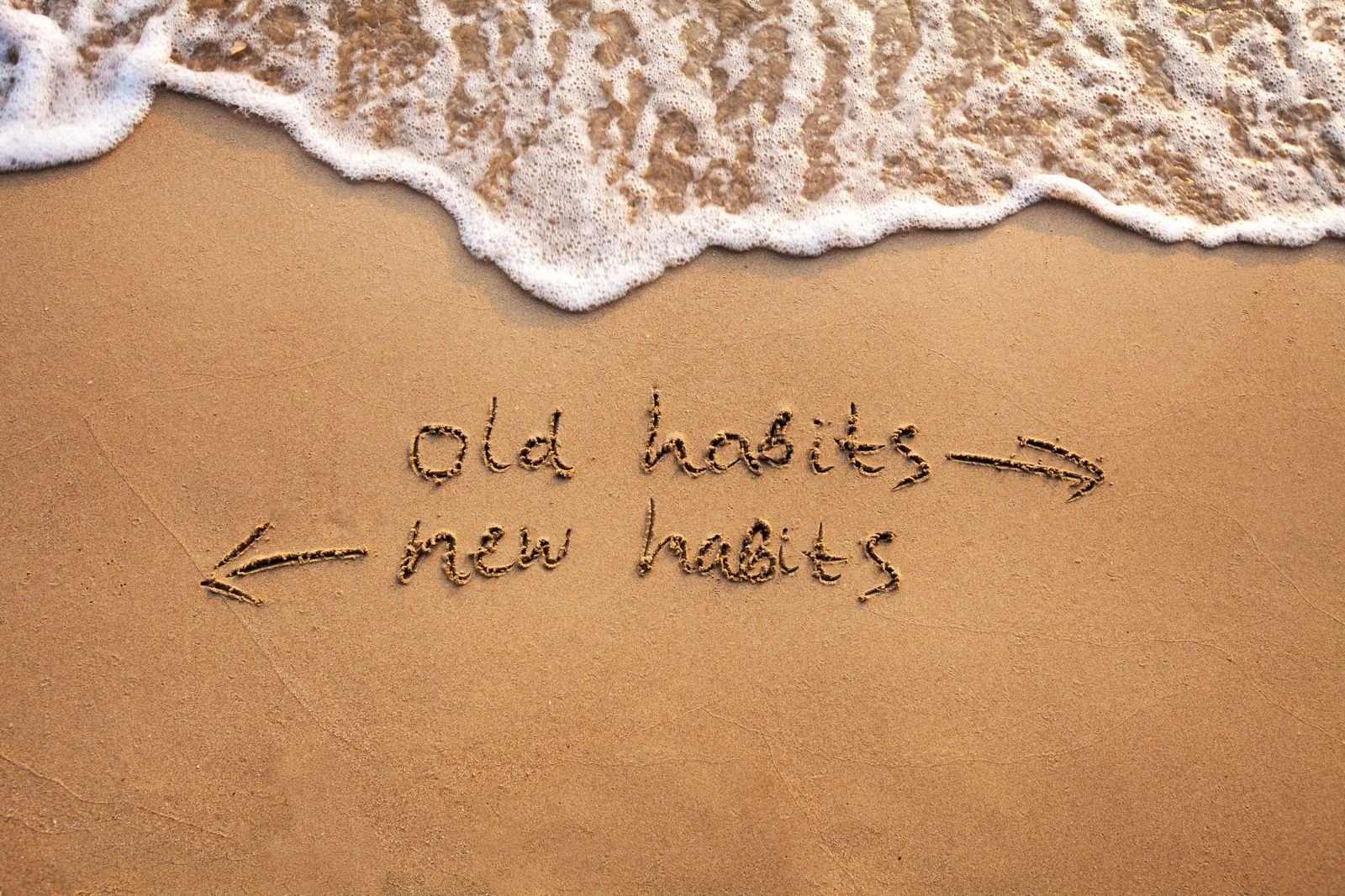 Writing in the sand on a beach comparing old habits to new habits, with an arrow pointing from 'old habits' to 'new habits'.