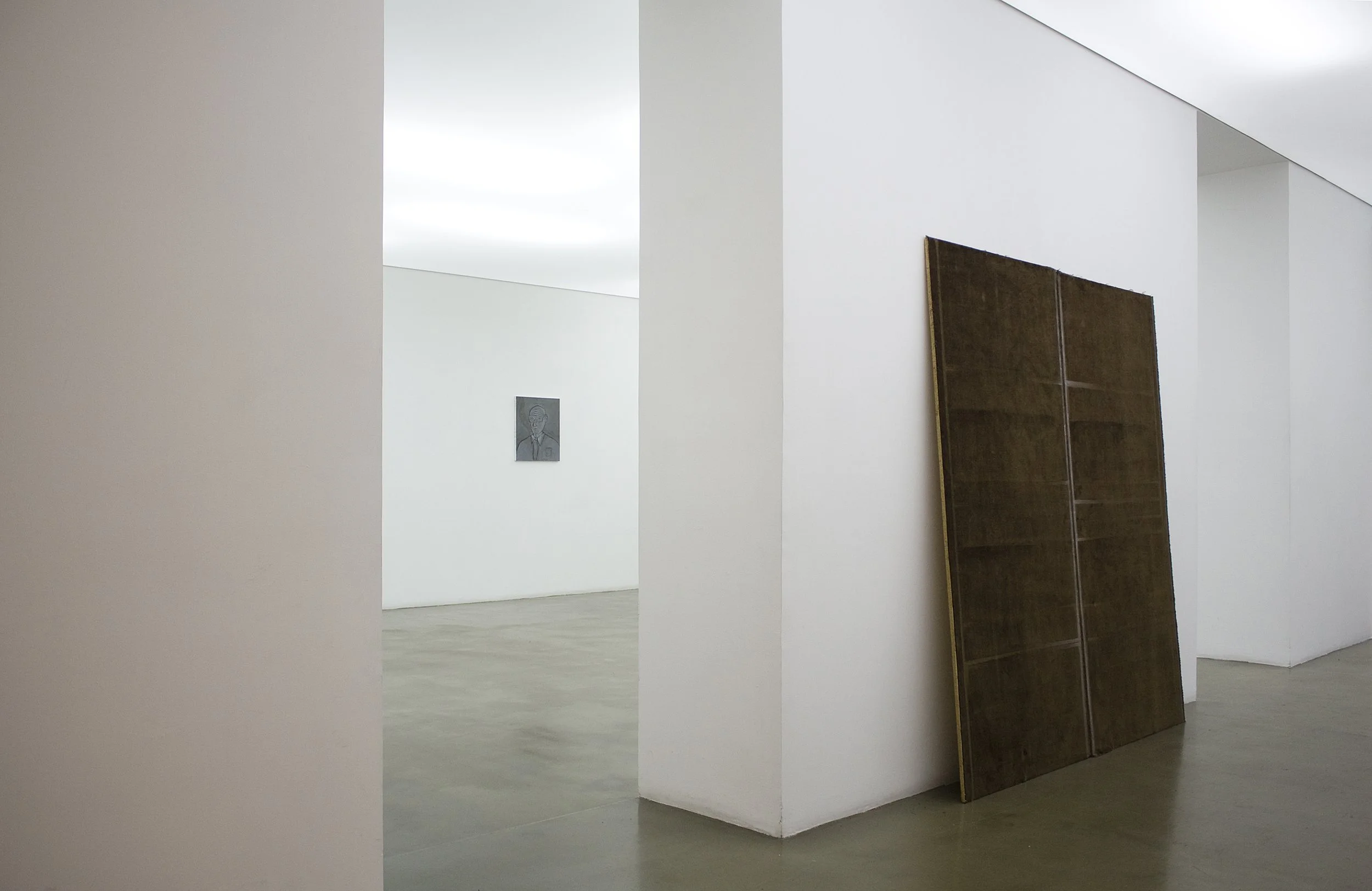 Interior of an art gallery with white walls, featuring a framed sketch of a person on the back wall and a partially folded wooden panel or furniture leaning against the wall.