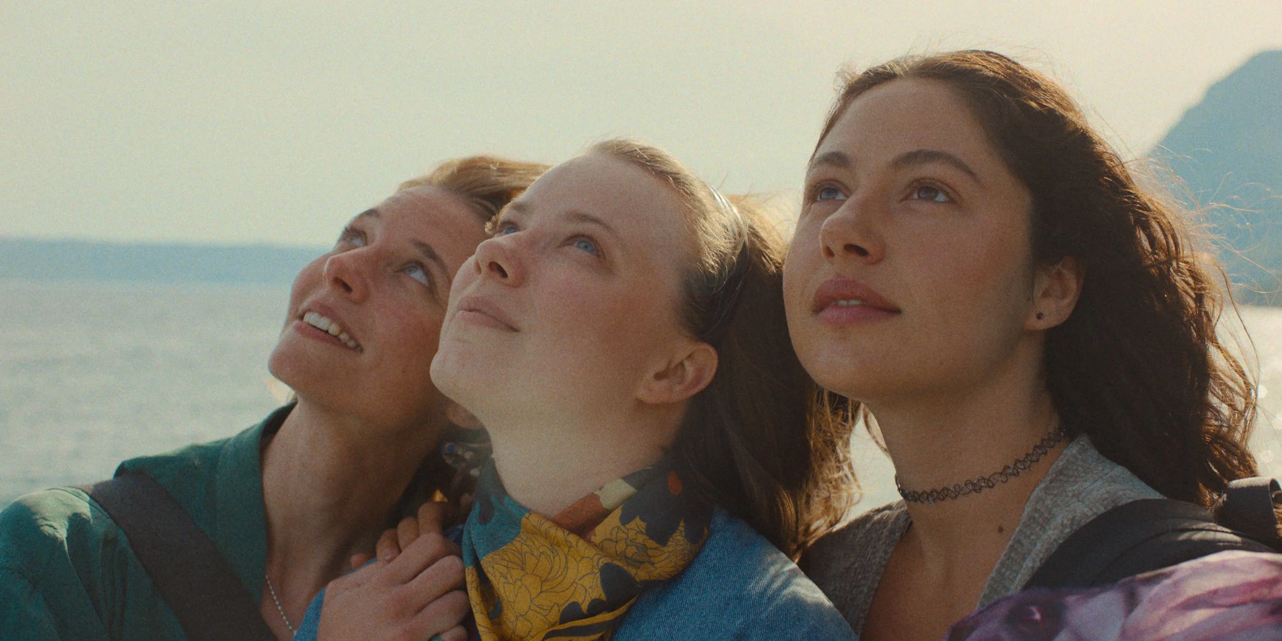 Three women with different hair colors and styles looking up with the sea and mountains in the background.