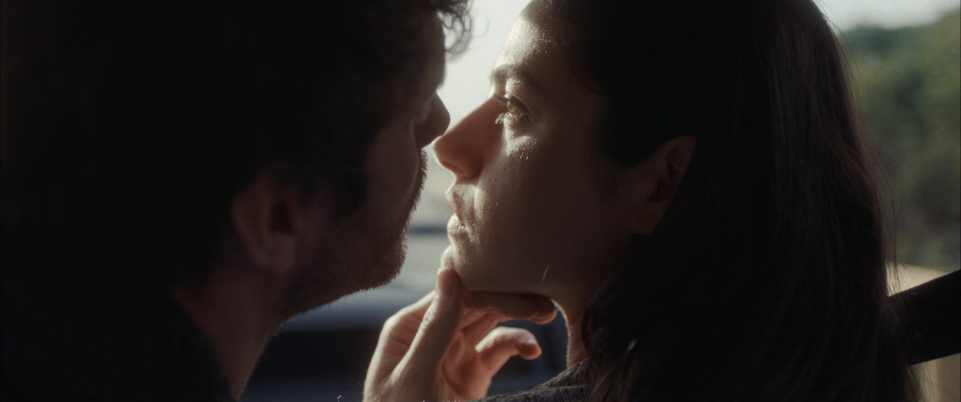 A close-up of two people with their faces inches apart inside a car, with a woman gently touching her chin, turned towards each other.