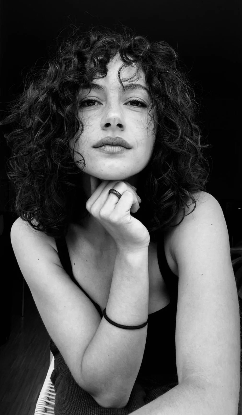 A black-and-white portrait of a woman with curly hair, resting her chin on her hand, looking into the camera.