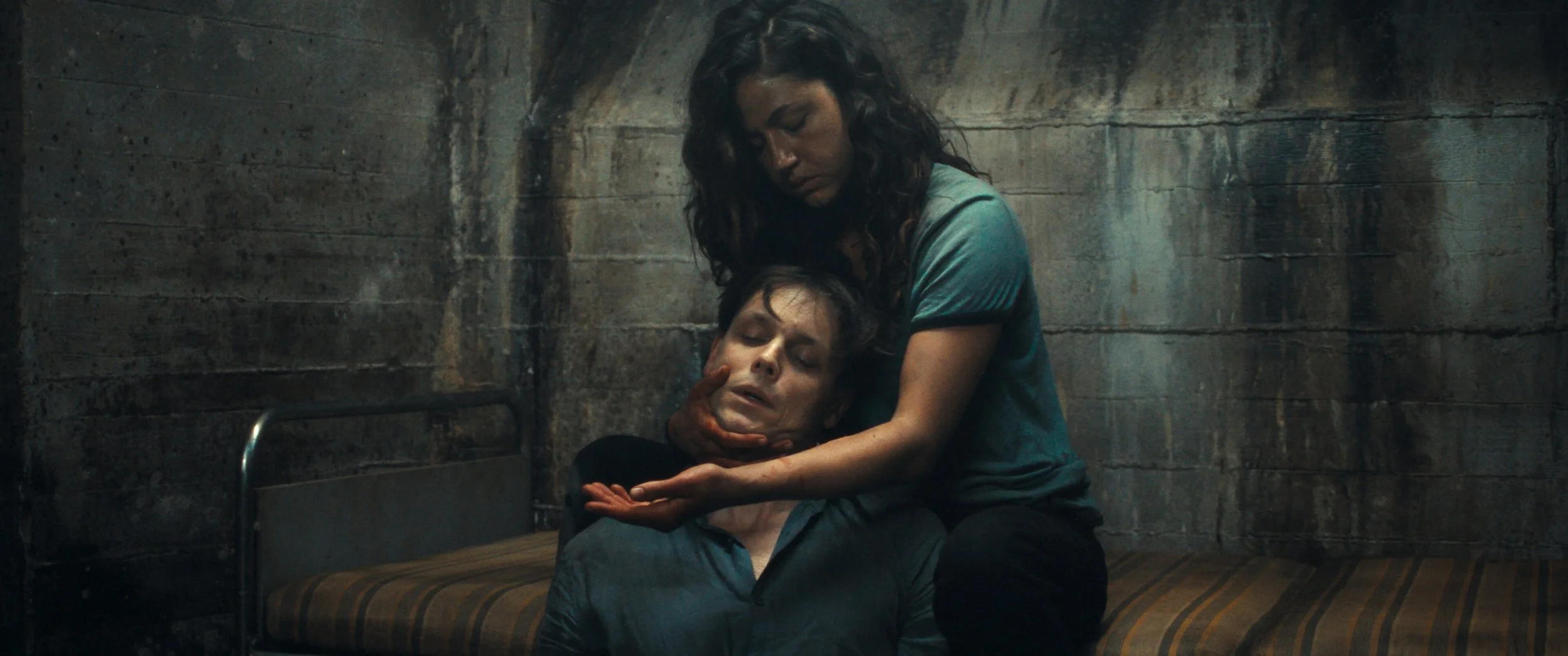 A woman comforts a man in a distressed state, sitting on a bed in a dimly lit, concrete-walled room.
