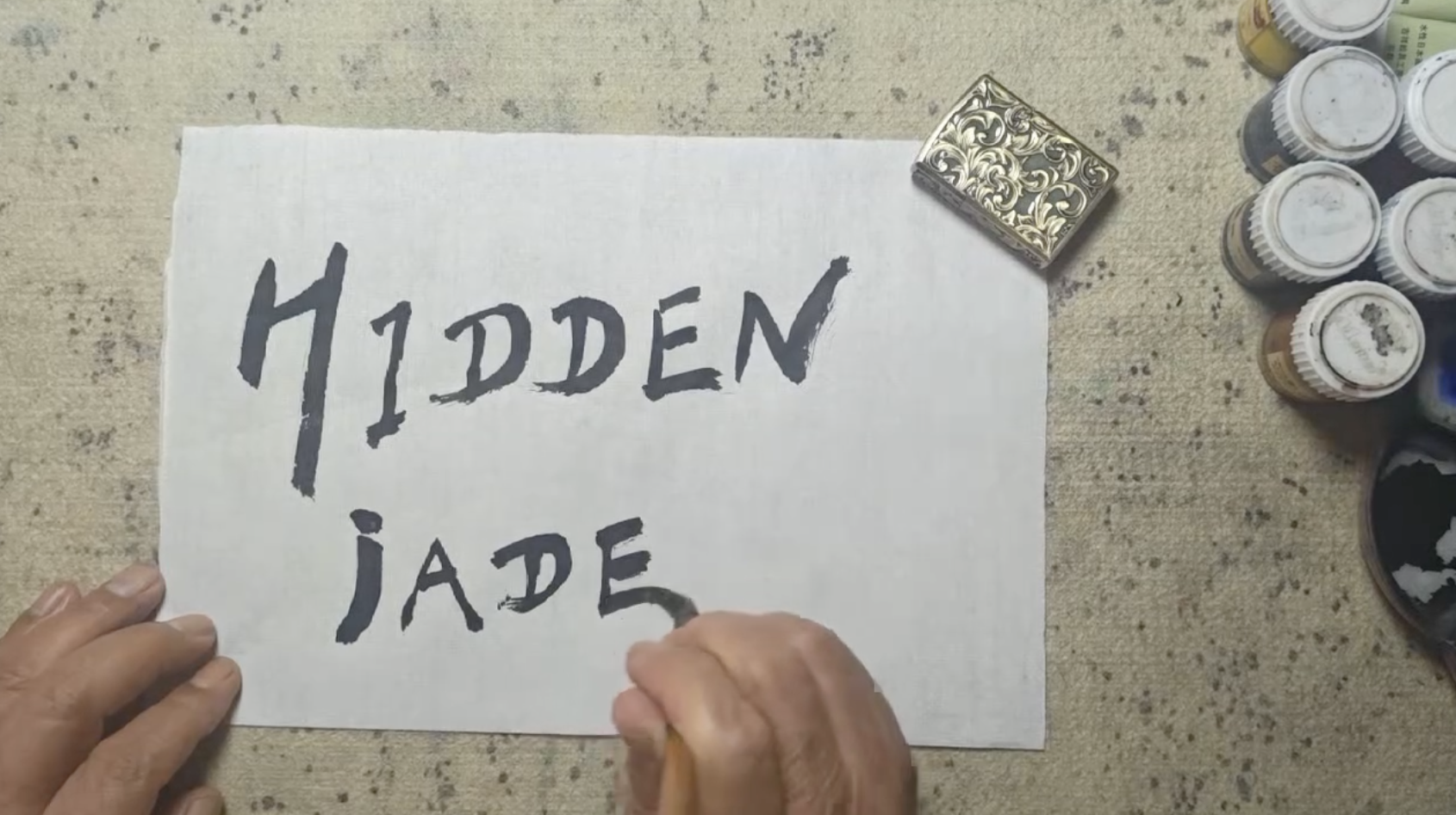 Artist writing the Hidden Jade Art Studio name in traditional brush calligraphy on handmade paper using black ink and a Chinese calligraphy brush.