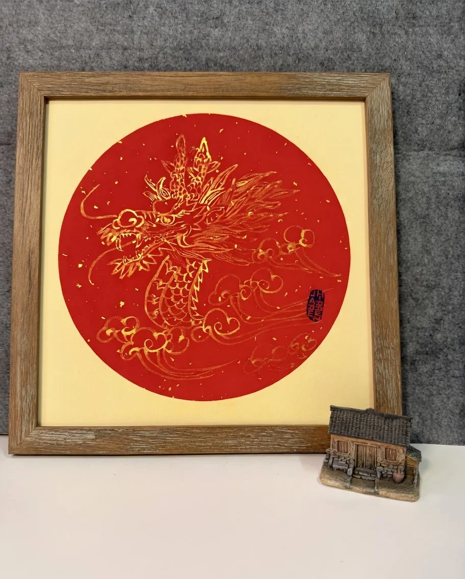 A framed picture of a gold dragon on a red circular background, with a small miniature house figurine placed in front of it on a table.