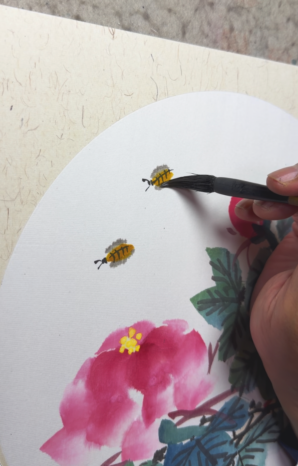 A person painting a watercolor picture of pink flowers, green leaves, and two bees in flight on a white canvas.