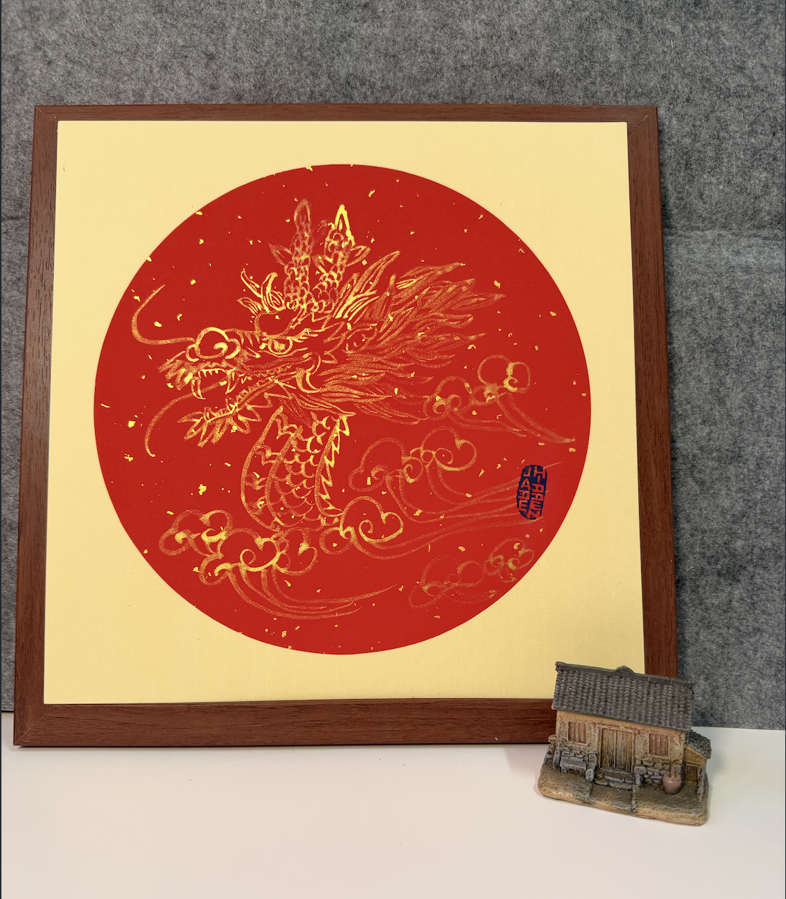 A framed Asian-style artwork featuring a gold-colored dragon on a red background, with Chinese characters in a blue seal. There is a small model of a traditional house or temple placed in front of the artwork.