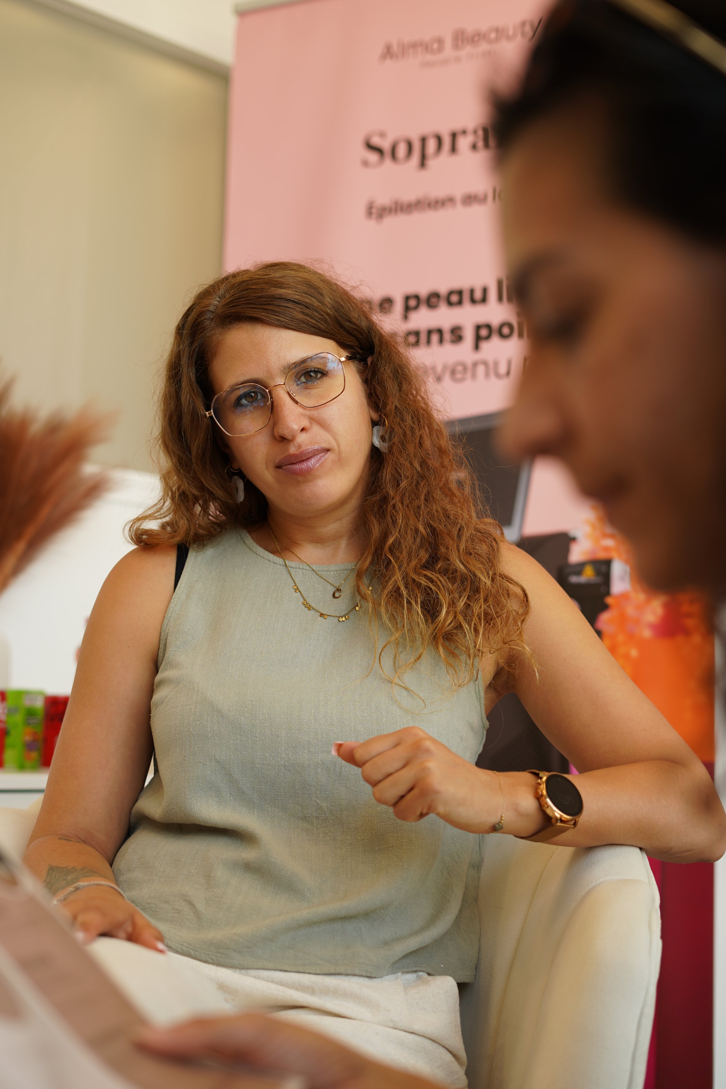Une femme brune avec des lunettes, portant un haut beige, assise dans une salle, regardant attentivement une autre personne qui est floue dans la partie droite de l'image.