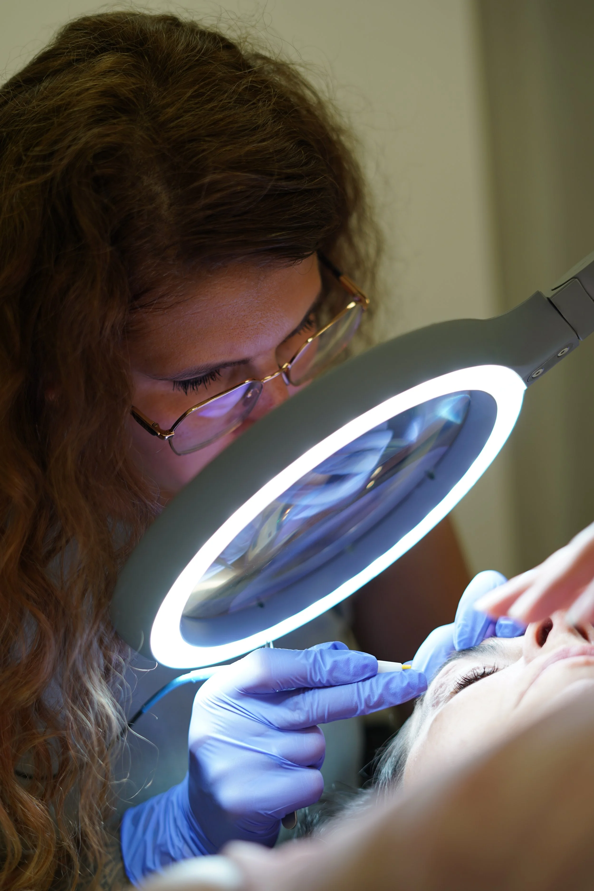 Une professionnelle en blouse blanche et lunettes effectue une thermolyse à une cliente à l'aide d'une loupe lumineuse. La scène se déroule dans un environnement professionnel