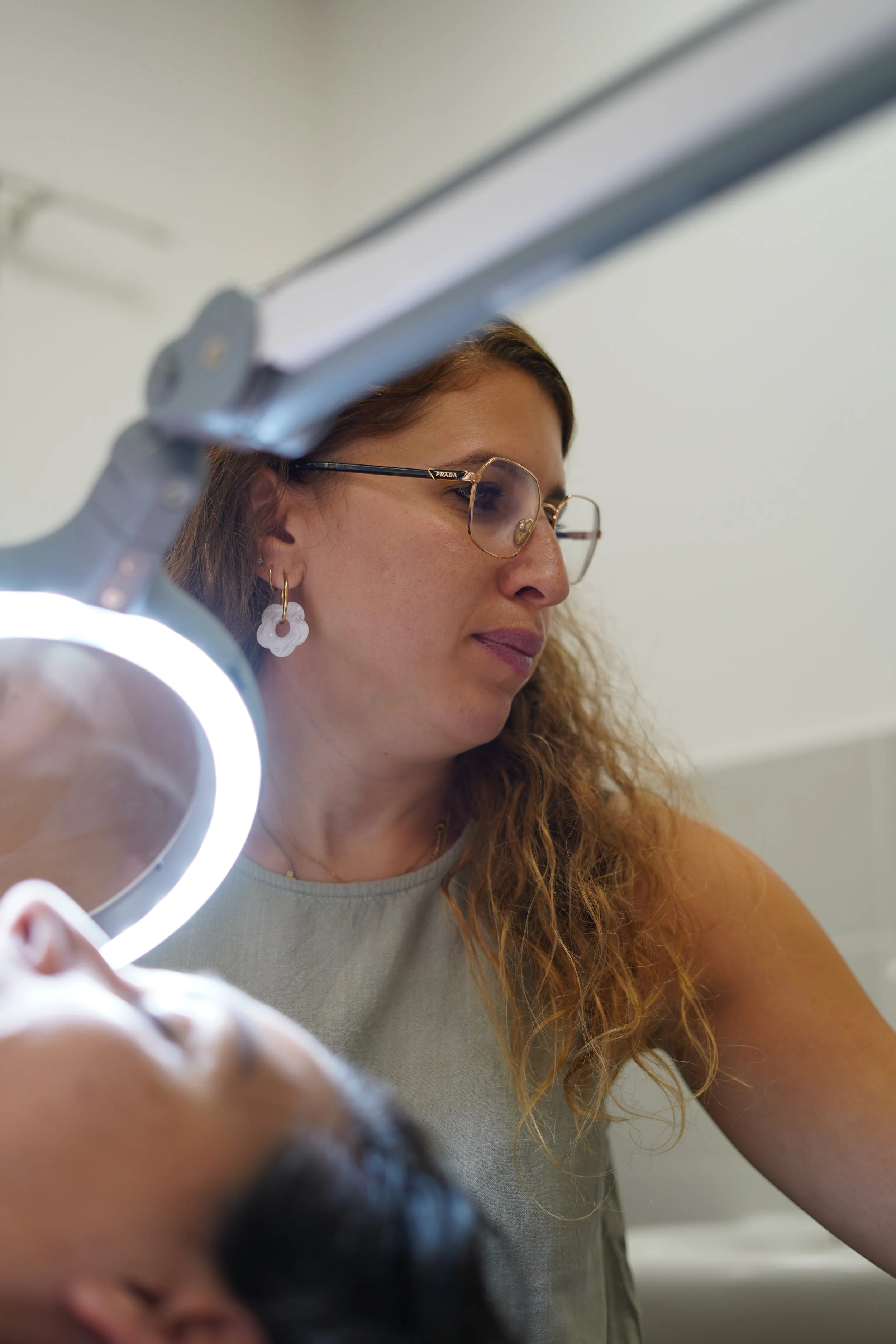 Une femme aux cheveux longs, portant des lunettes à monture dorée avec le logo Prada, et un earring blanc en forme de fleur, regarde vers la droite dans un environnement intérieur avec un mur blanc. La photo est prise en gros plan avec une lumière blanche circulaire dans le coin inférieur gauche.