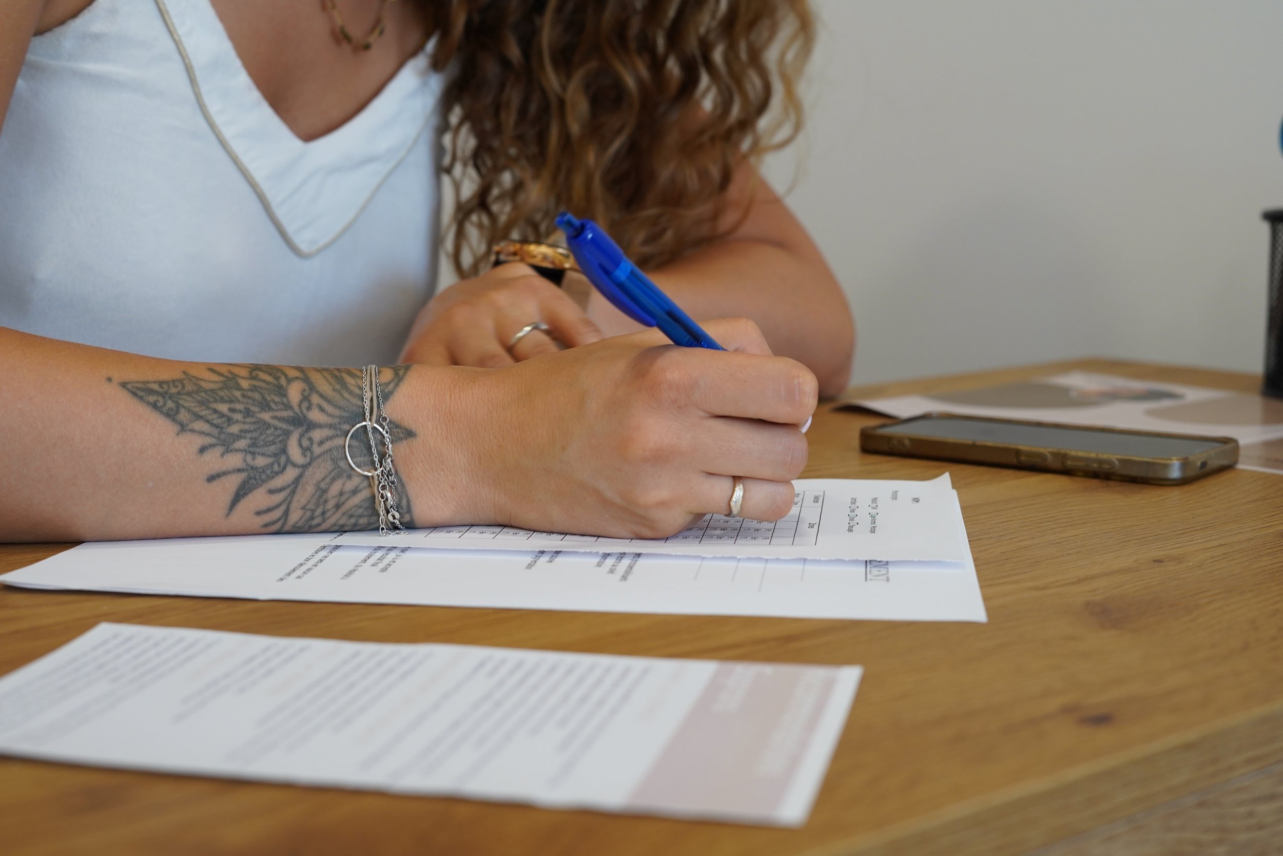 Une personne écrit sur un document avec un stylo bleu, posée à une table avec un smartphone et des papiers.