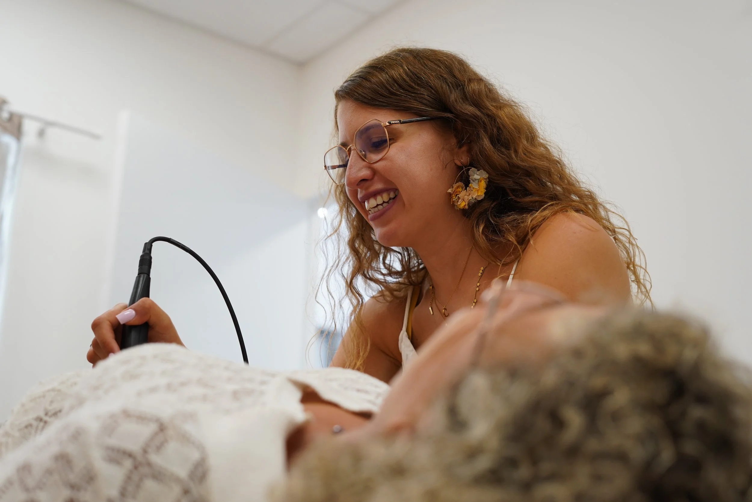 Une femme souriante portant des lunettes et des boucles d'oreilles en fleurs, tenant un stylo de luxoponture lors d'une conversation ou d'une interview, avec un fond blanc.