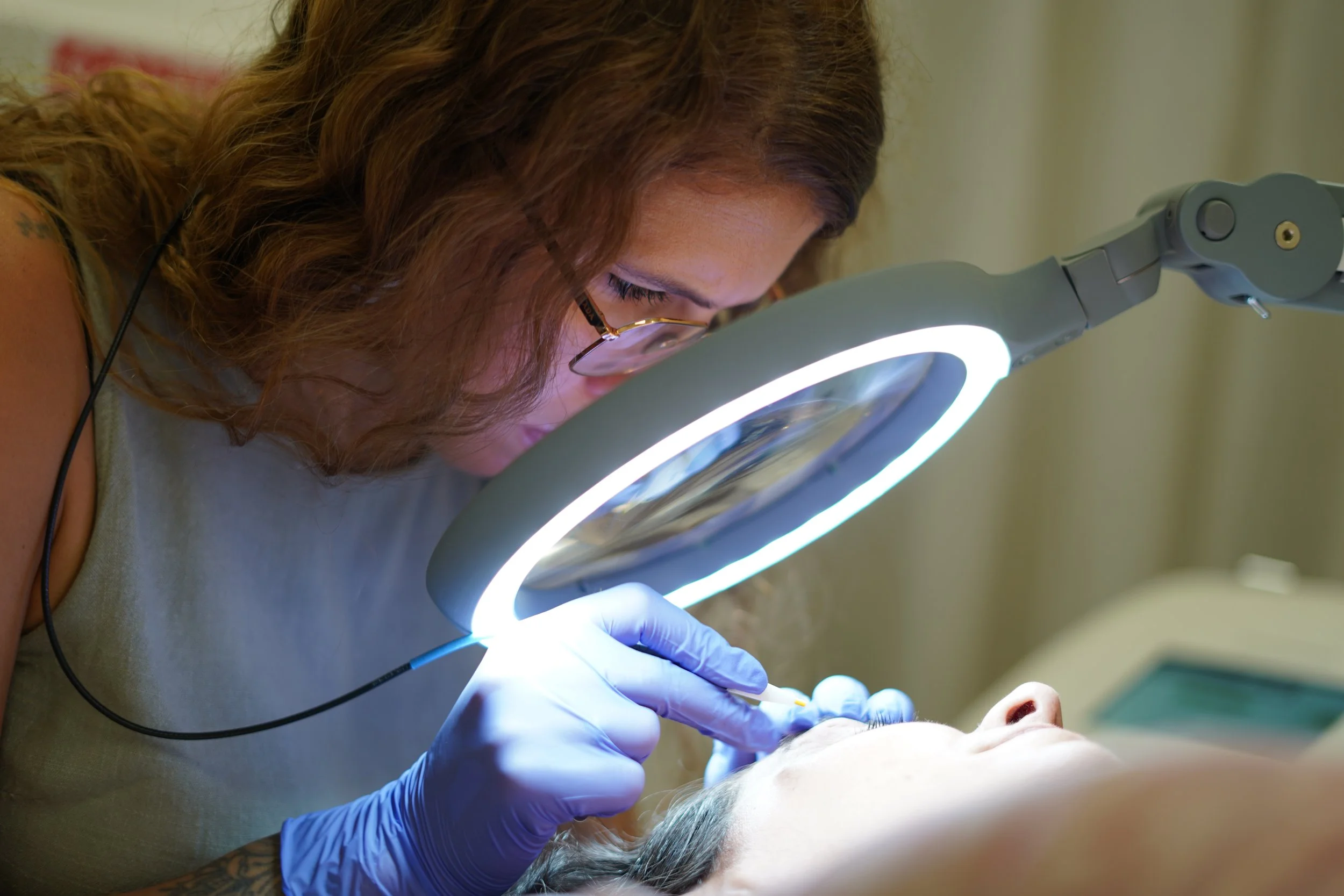 Une femme avec des cheveux bouclés, portant des lunettes, examine attentivement un patient à l'aide d'une loupe avec lumière intégrée