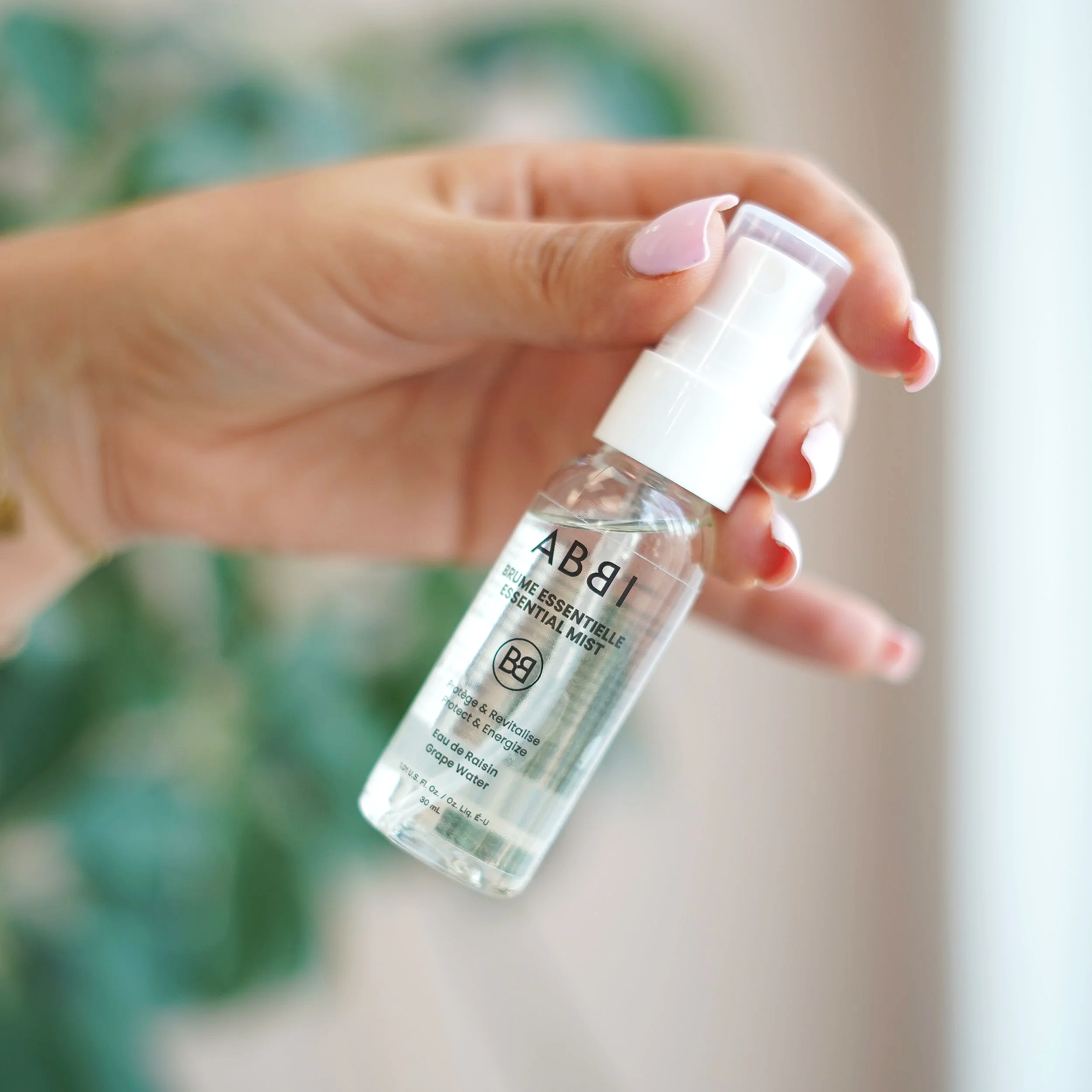 Un flacon de produit cosmétique ou de soin contenant un liquide clair, tenu par une main avec des ongles peints en rose. Le flacon porte une étiquette avec du texte en français.
