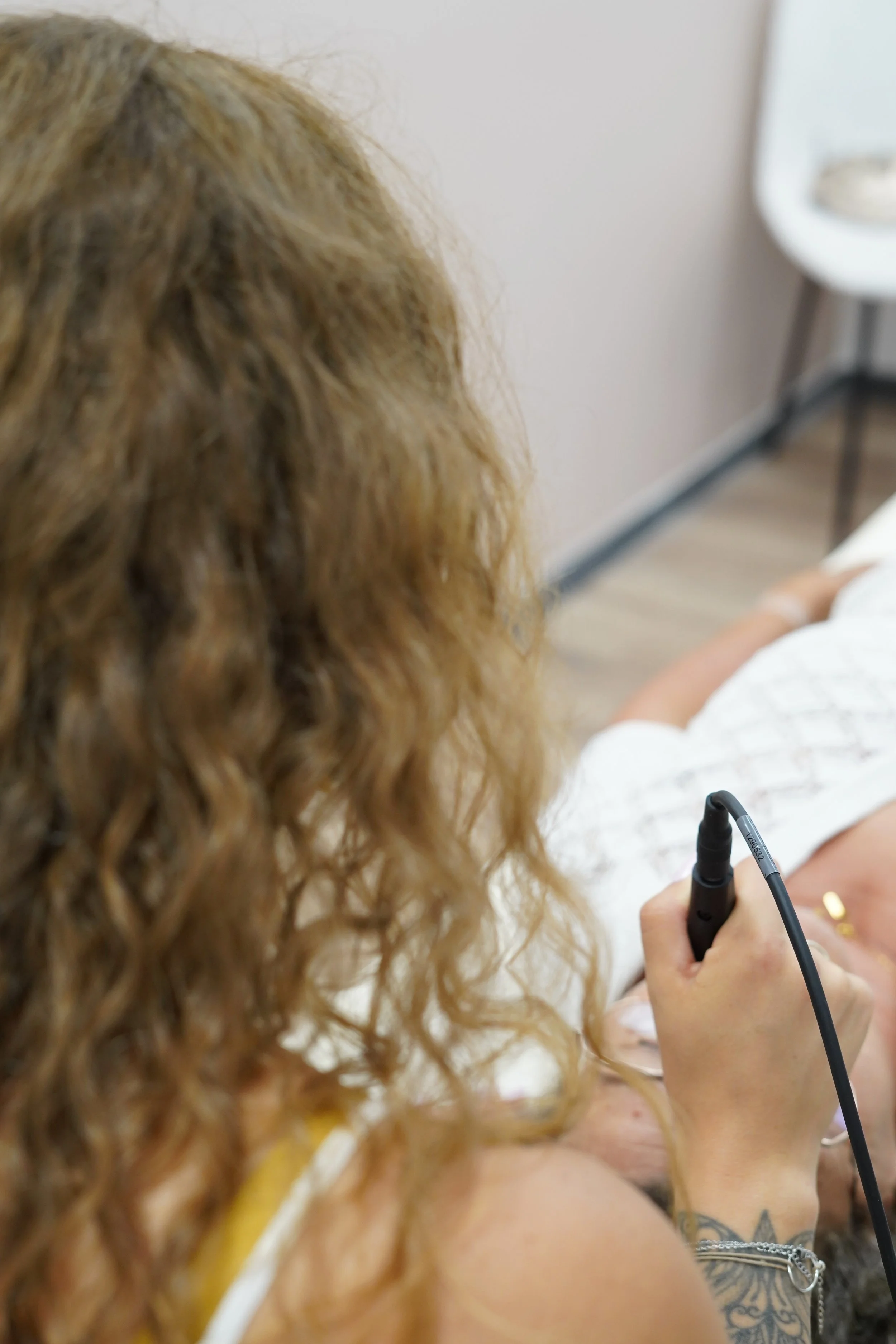 Une femme aux cheveux bouclés roux tient un stylo ou un outil électronique dans sa main, avec un tatouage visible sur son bras. L'arrière-plan montre un mur blanc et un meuble blanc.