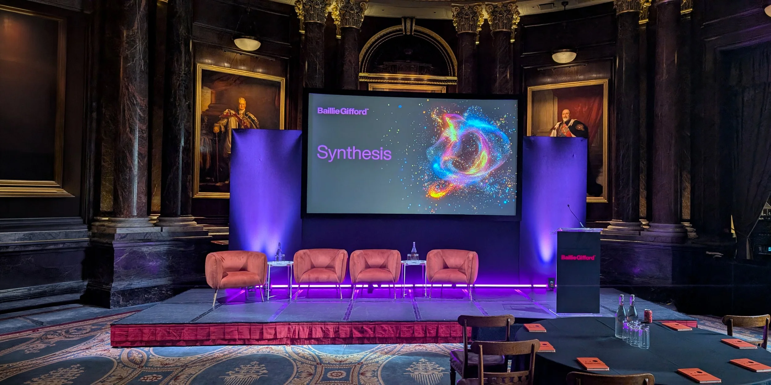 A conference room setup with a presentation screen that displays 'Baillie Gifford Synthesis' along with colorful graphics. There are four pink chairs on the stage with small tables and water bottles in front of each. The room has dark, ornate walls with portraits and columns, dim lighting, and a carpeted floor.