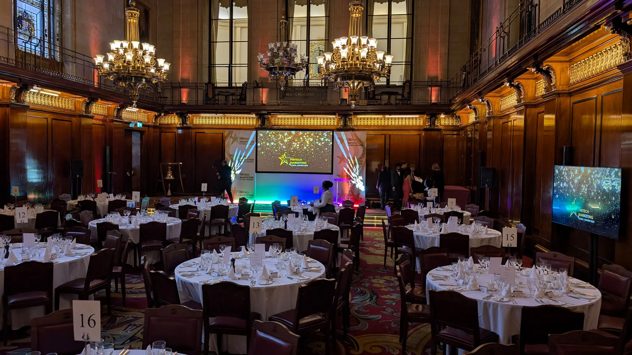 Elegant banquet hall with round tables set with white tablecloths, glassware, and silverware, a stage with large screens displaying 'Fintech Marketing Global Awards 2023,' ornate wood paneling, chandeliers, and high ceilings.