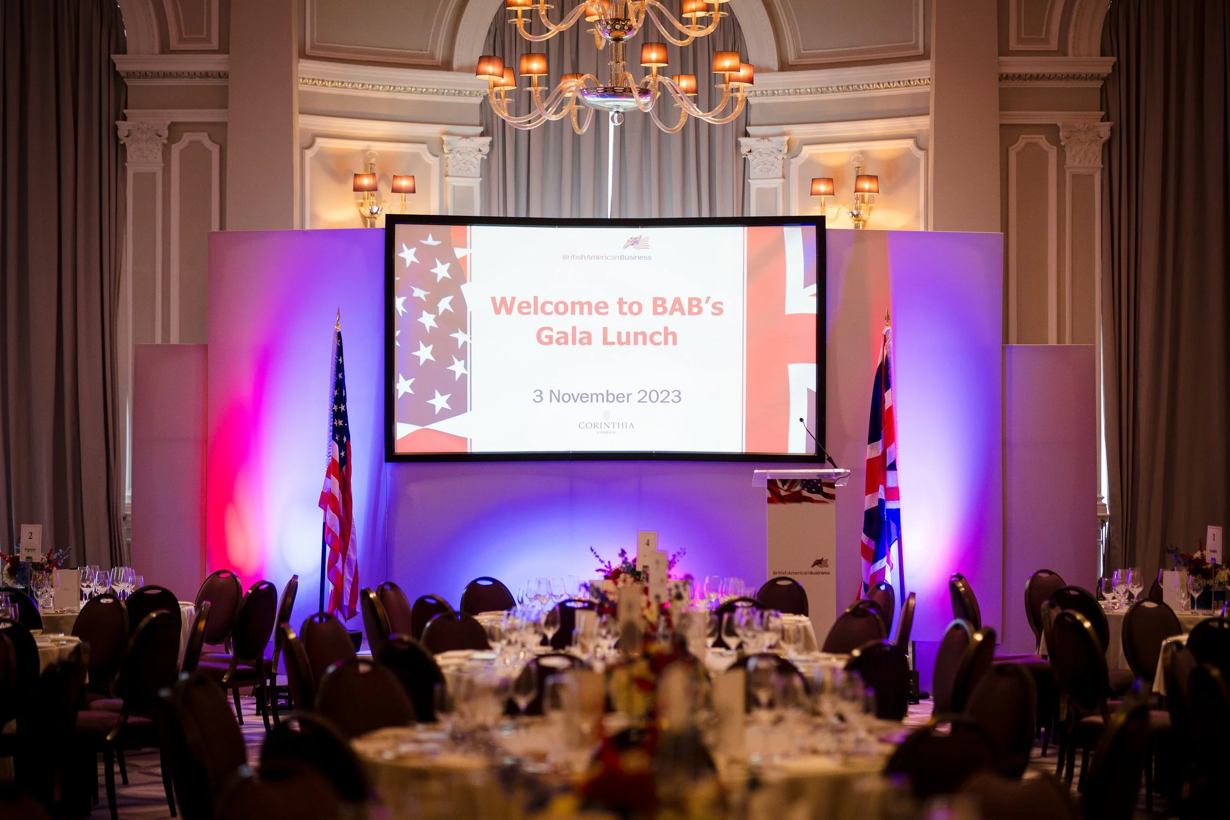 Ballroom decorated for a Gala Lunch event with tables set for dining, a large screen displaying a welcome message, flags, and a chandelier overhead.