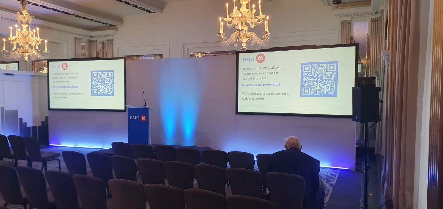 Conference room setup with two large screens displaying QR codes and instructions for accessing a poll, with rows of chairs and a few people seated, chandeliers hanging from the ceiling, and a lectern with a BMO logo.