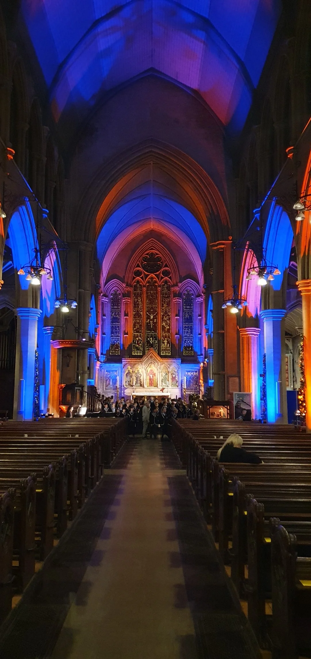 An illuminated church interior with colorful lighting in shades of blue, purple, and orange. The church has wooden pews, a high vaulted ceiling, stained glass windows, and an altar at the front where a choir is gathered.