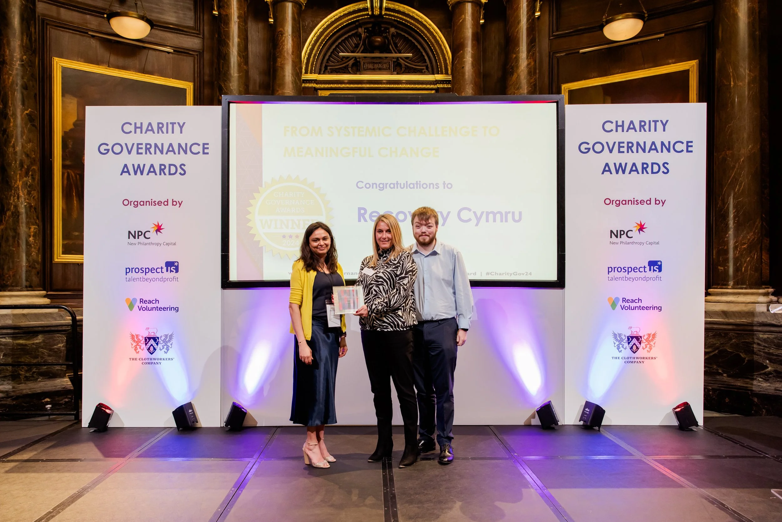 Three people standing on stage during an awards ceremony, with two women and one man, smiling at the camera. The background features a screen displaying text about charity and governance awards, flanked by two large white banners with logos and sponsors. The setting appears to be a formal event in a grand hall with decorative wood paneling and lighting.