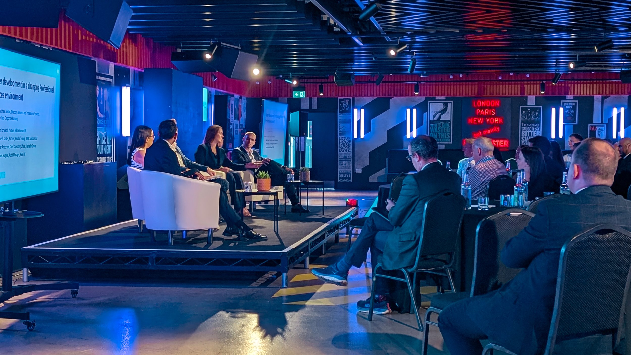 A panel discussion with four speakers on stage in front of an audience in a modern, vibrant conference room decorated with neon lights and posters. Audience members are seated, listening attentively.
