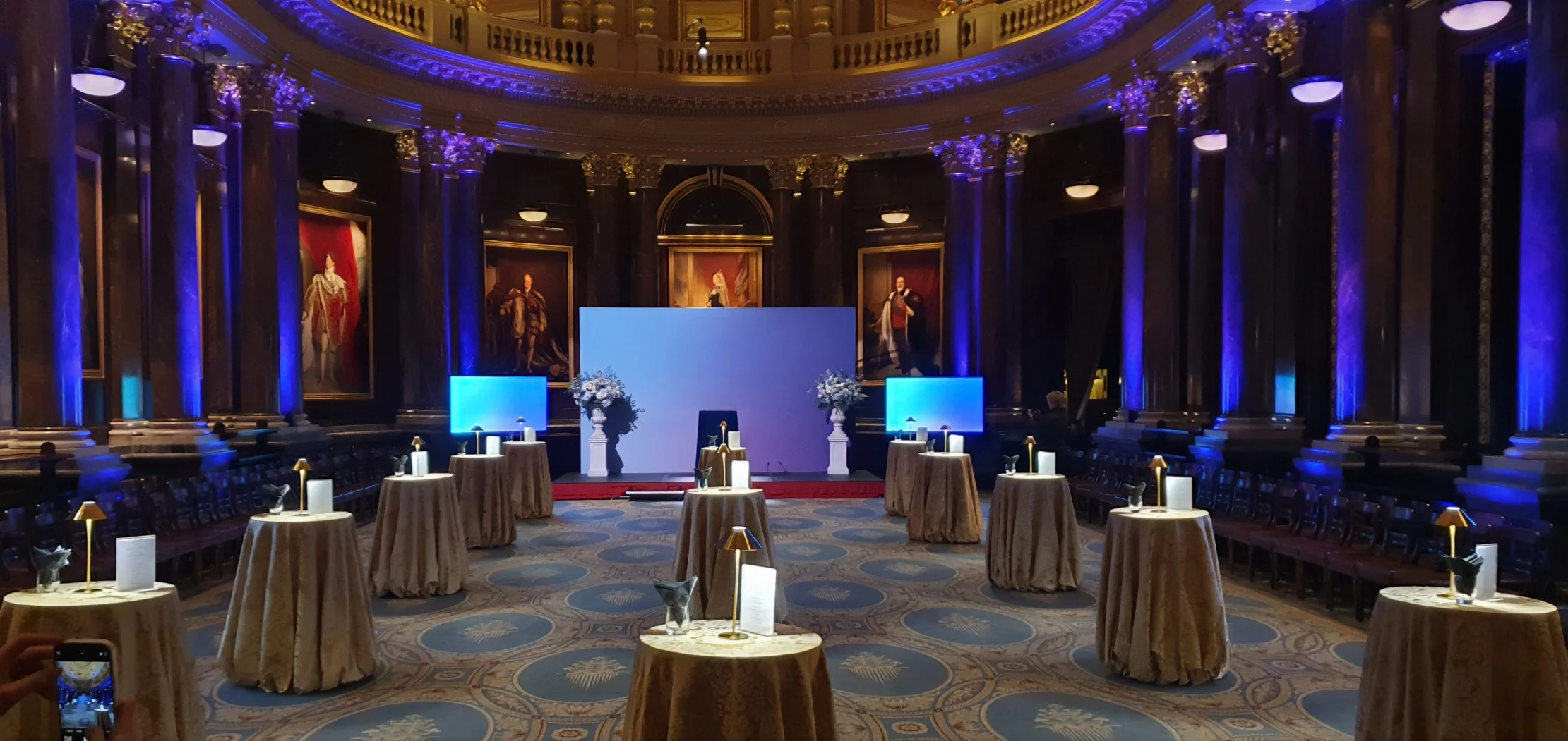 Elegant event space with round tables draped in gold tablecloths, each with a small lamp and flower arrangement, set in front of a stage with a large blank screen, flanked by two large floral arrangements. The room features tall, dark columns, ornate gold accents, grand portraits on the walls, and blue lighting creating a sophisticated atmosphere.