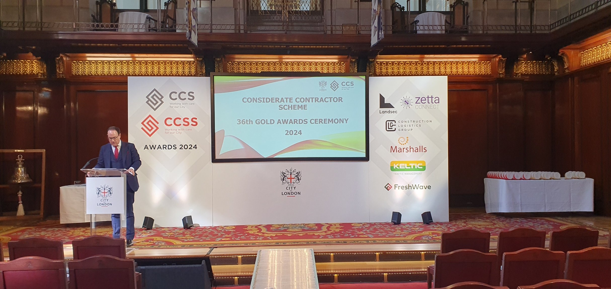 A man in a suit and glasses stands behind a lectern with the City of London emblem, speaking at a conference or awards ceremony in a grand wooden hall. There is a large screen behind him displaying the event title "Considerate Contractor Scheme 36th Gold Awards Ceremony 2024". On either side of the screen are banners with various company logos, including CCS, CCSS, Landsec, Zetta Connect, Construction Logistics Group, Marshalls, Keltic Traffic Management, and FreshWave. To the right, a table holds numerous rolled-up certificates or awards.