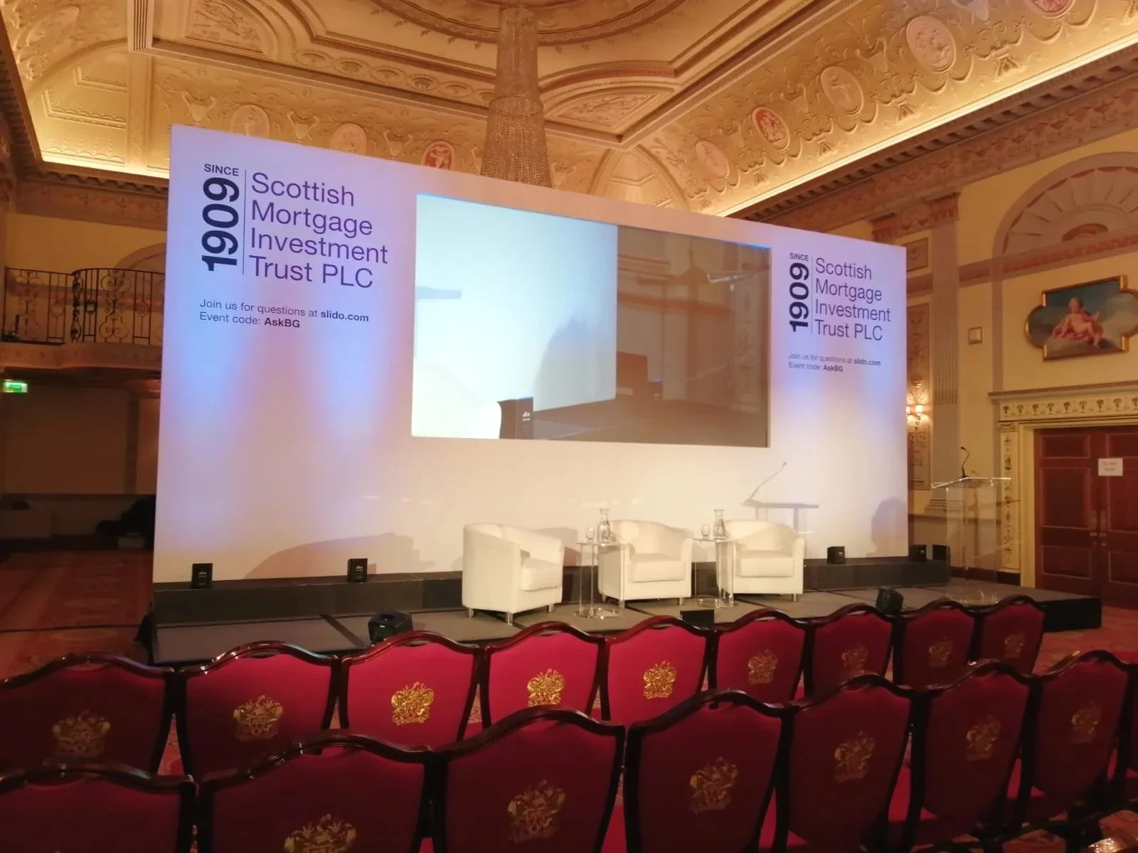 The image shows a stage set up for a presentation or panel discussion, with a large screen on a white backdrop displaying information about the Scottish Mortgage Investment Trust PLC, established in 1909. The screen also has details about joining questions on a website. In front of the stage, there are three white armchairs and a small round table with water bottles and glasses. The room has ornate gold and cream-colored decor with chandeliers, decorative moldings, and red upholstered chairs in the audience area.
