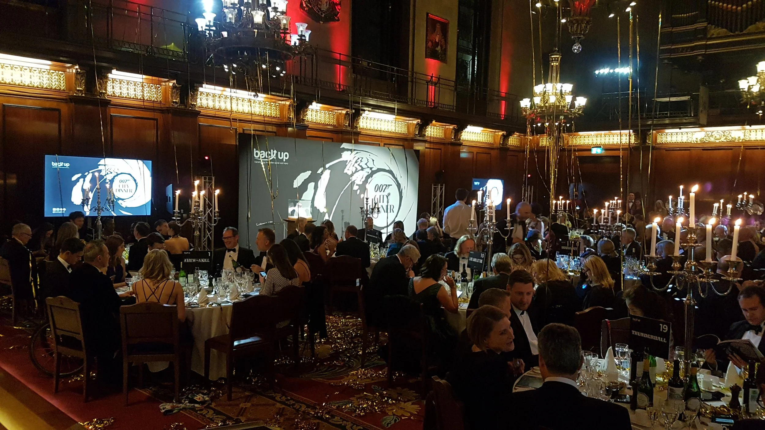 A formal banquet in a large, ornate ballroom decorated with chandeliers, candelabras, and gold accents. Guests in tuxedos and evening gowns sit at round tables, enjoying dinner and conversation. A large screen displays the James Bond theme, and people are socializing in a sophisticated setting.