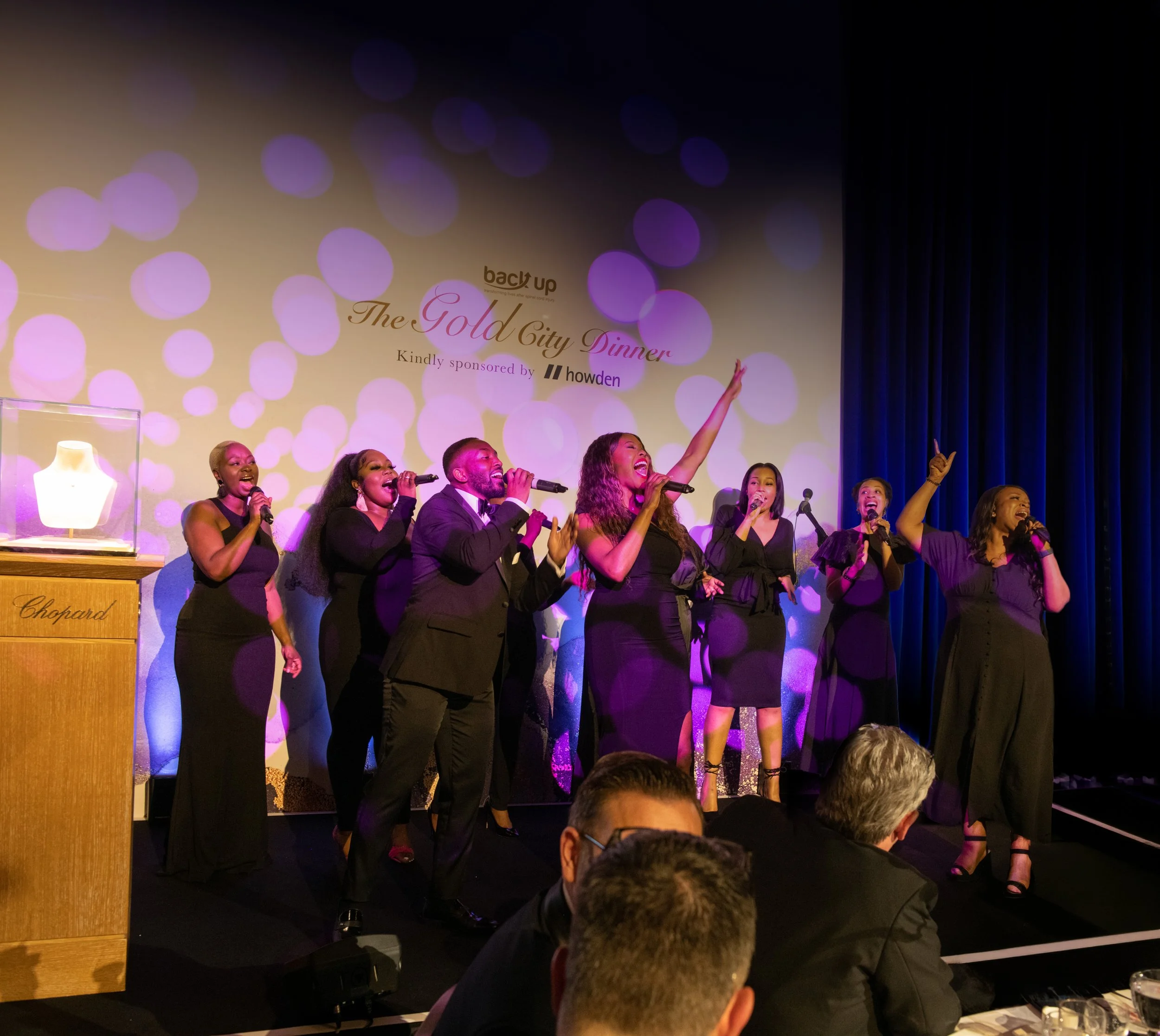 A group of people singing on stage at an event called back up The Gold City Dinner, with purple and pink bokeh lights in the background.