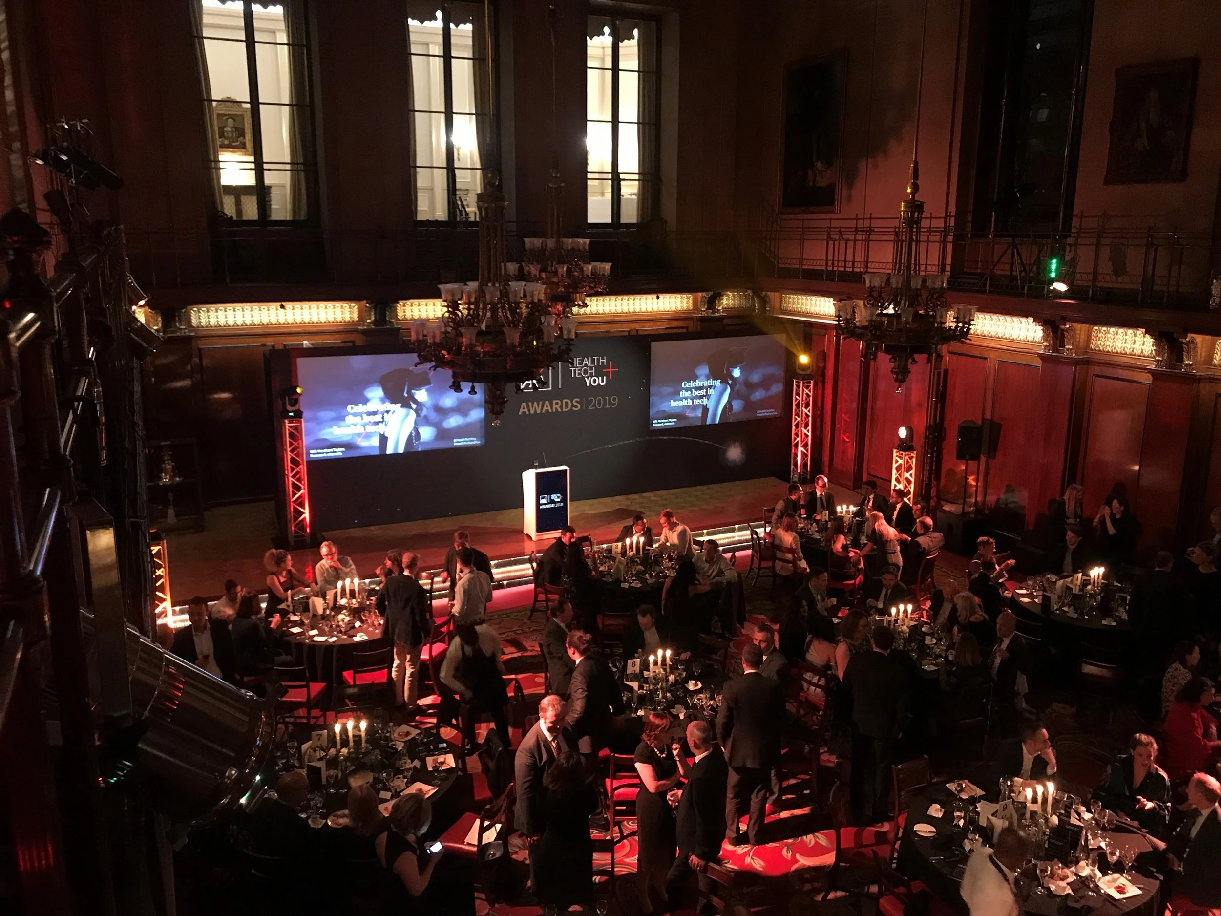 A large elegant ballroom set up for a formal event with round tables, candles, and guests dining. A stage with screens displays 'Health Tech You Awards 2019'.
