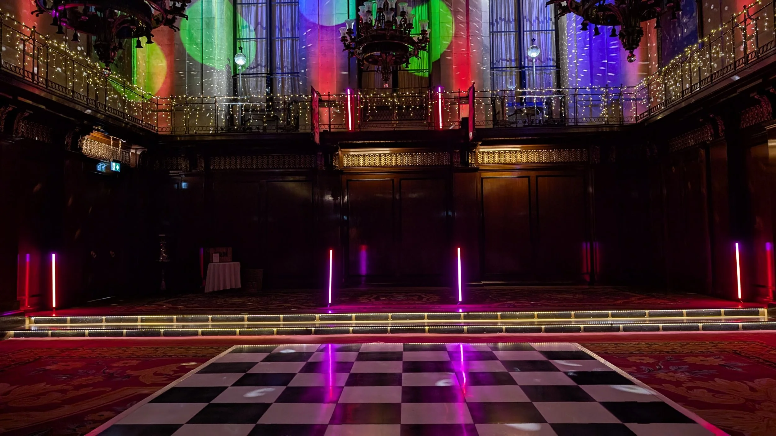 Empty dance floor with checkered black and white tiles, colorful stage lighting, and elaborate chandeliers in a grand hall decorated with string lights and vibrant projected patterns on the walls.