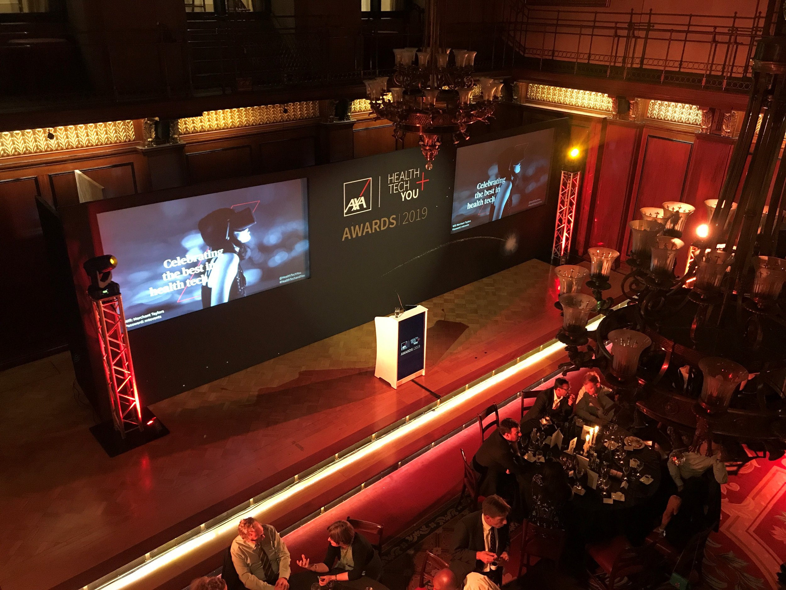 An indoor event with a stage displaying the AAAX Health Tech You Awards 2019 logo and a large screen showing a person with pink hair. The room has ornate wood paneling, chandeliers, and people seated at tables and mingling.
