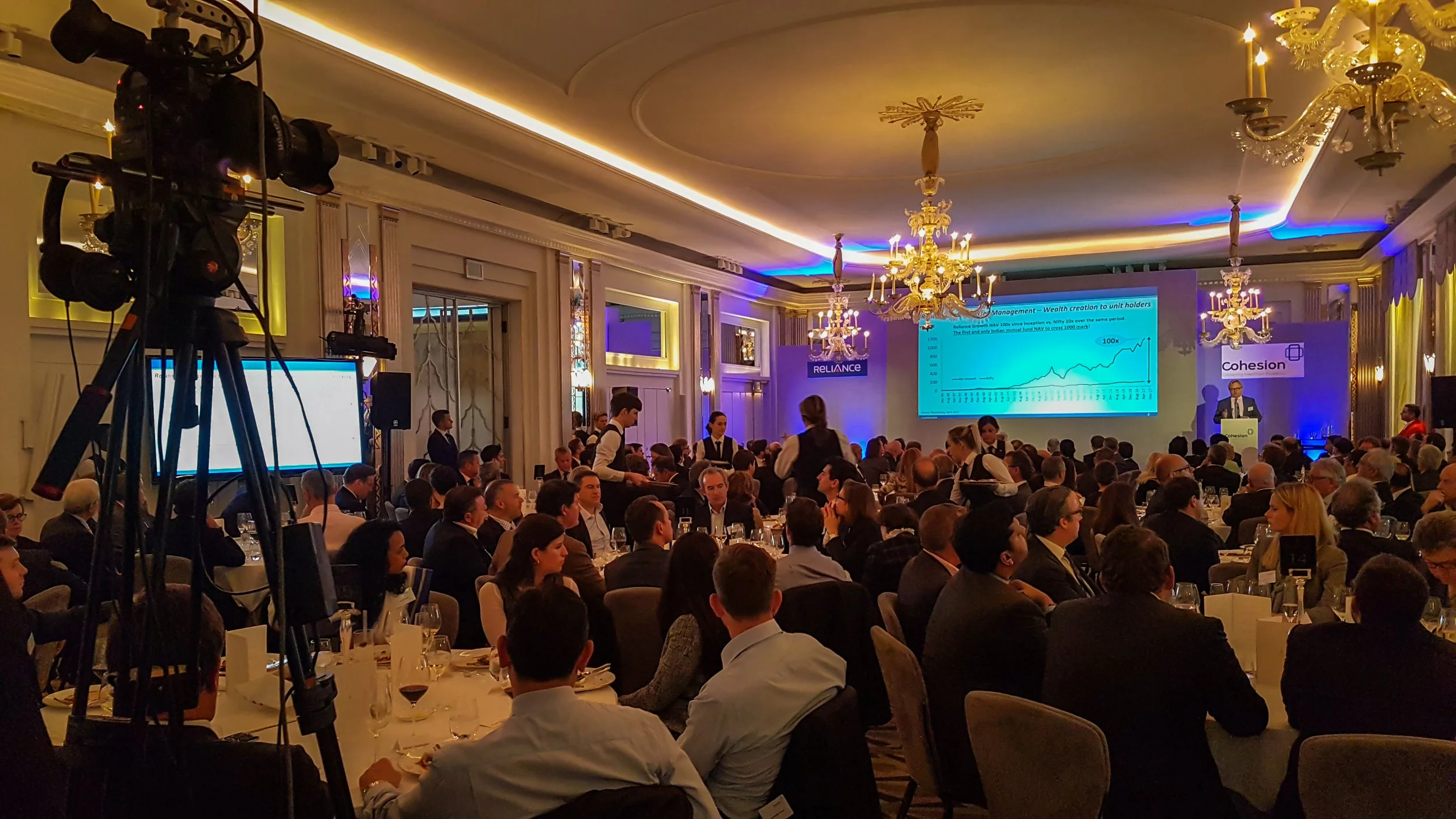 A large conference room filled with many people sitting at tables, attending a presentation with slides projected onto a large screen. There are chandeliers hanging from the ceiling and staff members serving at the tables.