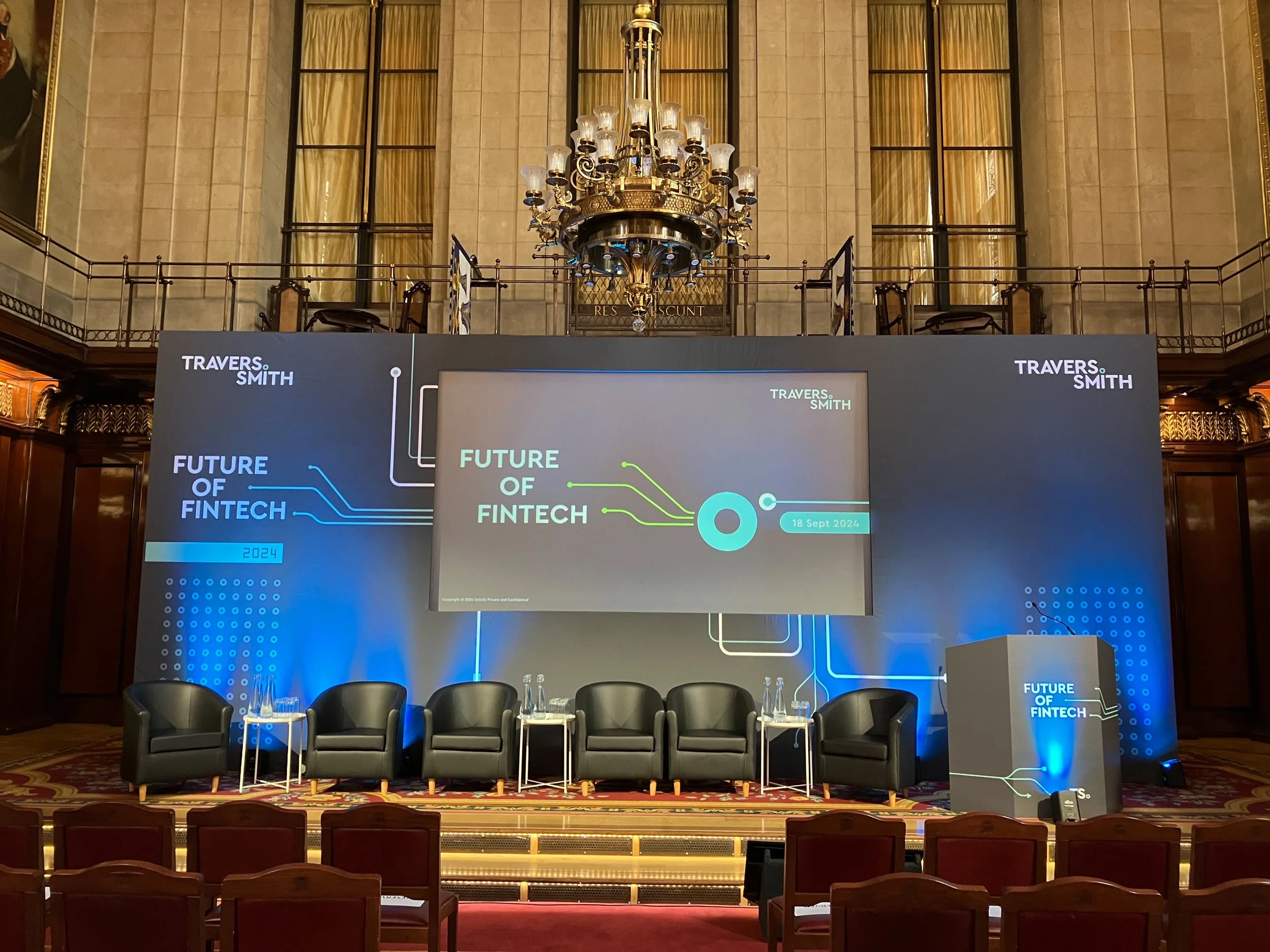 A stage set up for a conference called 'Future of Fintech' with a large digital screen, five black chairs, and two small tables with water bottles and glasses, all within a historic hall with a high ceiling, ornate walls, and a grand chandelier.